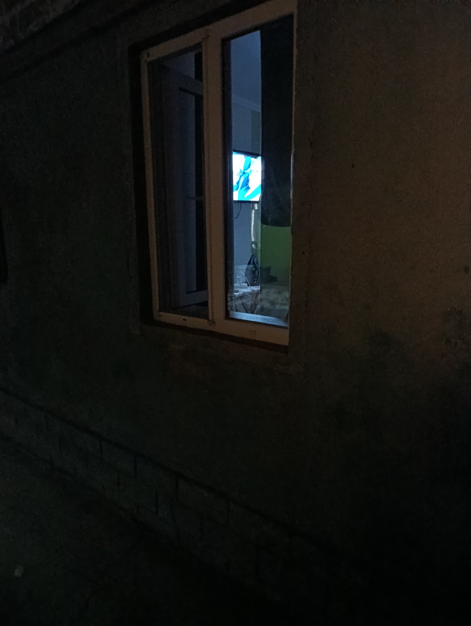 The image depicts an exterior view of a building at night in Roghi, Moldova. A window is visible, with light emanating from within. Through the window, a television screen is illuminated, displaying a bright, abstract image with shades of blue and white. The exterior walls of the building appear rough and dark, with some texture visible. A darkened foreground suggests a street or pathway. The scene is dimly lit, indicating nighttime conditions.