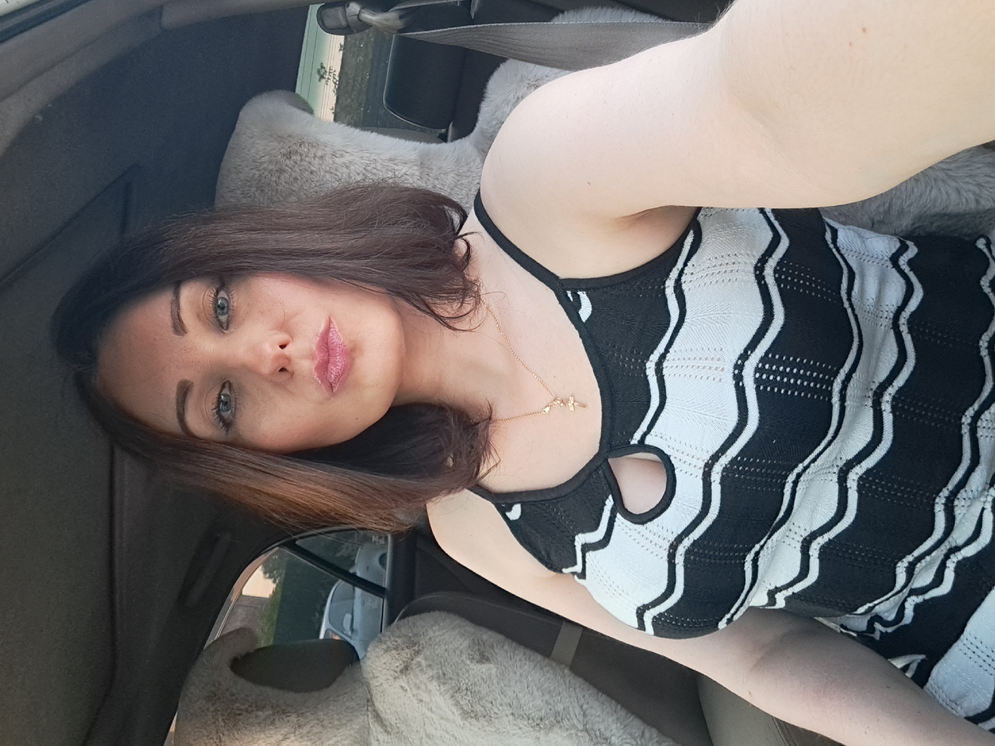 A woman with dark hair, fair skin, and blue eyes is sitting in the passenger seat of a car. She is wearing a black and white striped top and a gold necklace with a cross pendant. The woman is looking at the camera and pursing her lips. In the background, two large stuffed animals, possibly teddy bears, are visible in the back seats of the car. Outside the car windows, a street scene with a car and buildings can be seen. The lighting suggests it is daytime.