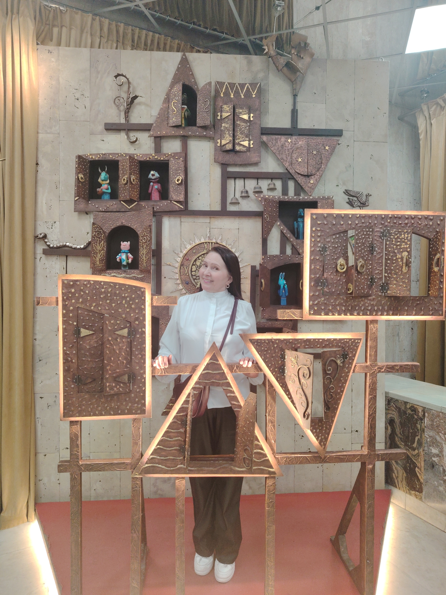 A full shot captures a woman standing behind a geometric art installation. The woman has dark hair and is wearing a white blouse, brown trousers, and white shoes. She has a purse slung over her shoulder. The art installation is made of copper-colored metal, with various shapes and designs. Behind the installation is a wall with a decorative pattern, and there are curtains on either side. The location appears to be inside a studio or exhibition space. The city and country are unknown.