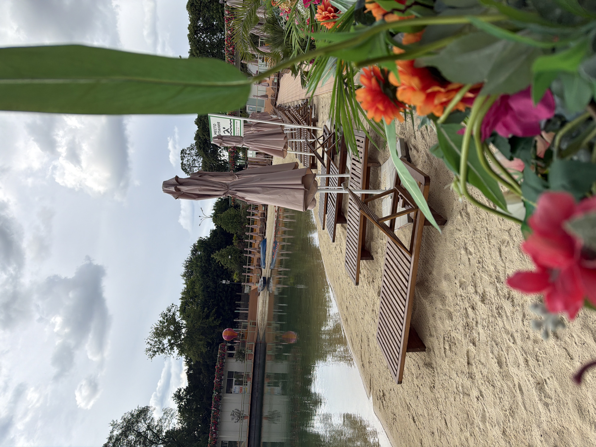 This image depicts a beach scene in Moscow, Russia. In the foreground, there are colorful artificial flowers, including red, pink, and orange ones, partially obscuring the view. Behind these, several wooden beach chairs and closed brown umbrellas line the sandy shore of a body of water. A sign, partially obscured, is also visible. The water reflects the sky and surrounding trees. In the background, there is a building decorated with flowers and what appears to be some canoes or kayaks are on the shore. Above, the sky is filled with white clouds.