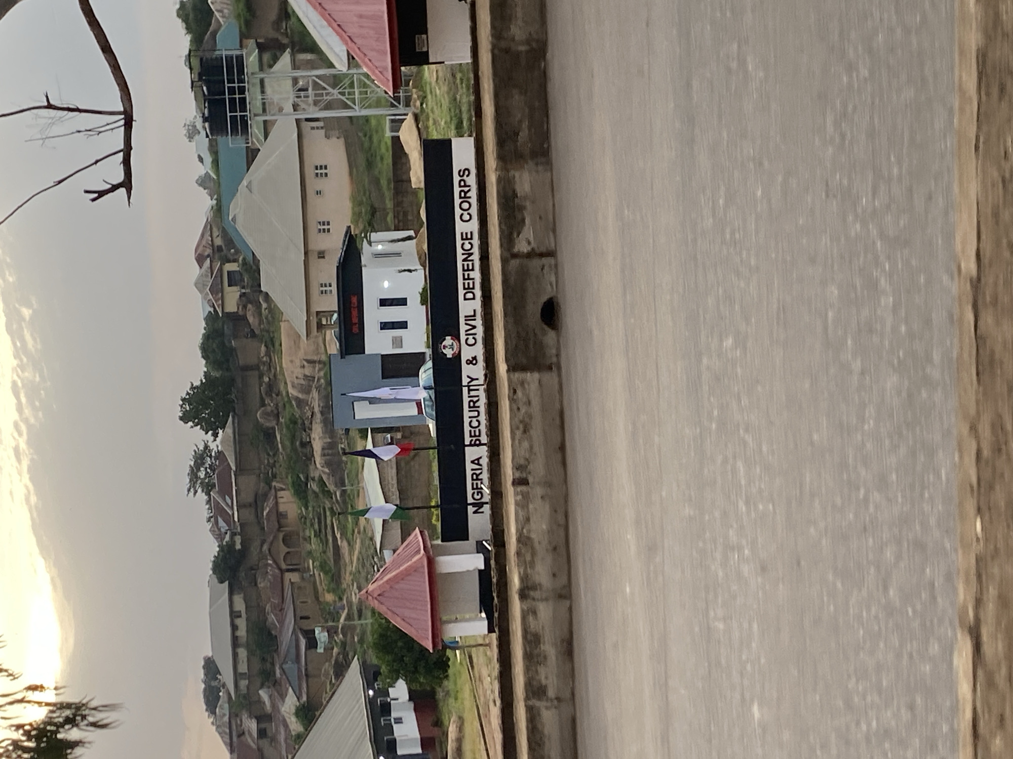 The image depicts an outdoor scene in Barakin Lerawi, Nigeria, on a cloudy day. In the foreground, a concrete barrier with a drainage hole separates the road from the background. Behind the barrier, a black sign with white lettering reads "NIGERIA SECURITY & CIVIL DEFENCE CORPS." To the left of the sign is a small white structure with a red roof, resembling a guard post. Three flags are visible behind the sign: a green and white flag, a white, red, and blue striped flag, and a white flag with blue markings.
In the middle ground, several buildings are visible, including a white building with a dark roof and a structure with a water tank on top. These buildings are nestled among rocky terrain and vegetation. The background reveals a hillside dotted with more houses, some with corrugated metal roofs. The overall environment appears to be a semi-urban or rural setting. The sun is partially visible through the clouds, suggesting it is either early morning or late afternoon. No people are clearly identifiable in the image.