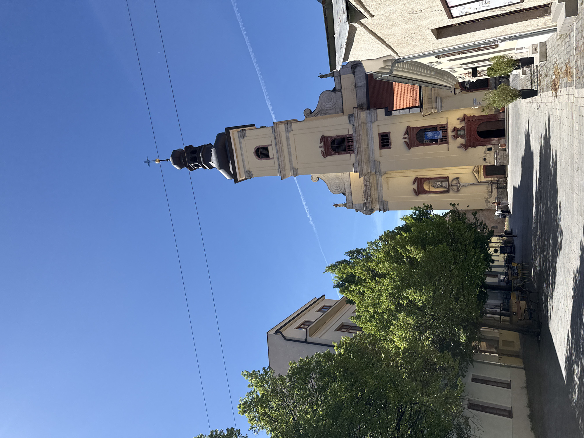 The image captures the Tower of St. Nicholas Cathedral in Trnava, Slovakia, framed against a clear blue sky. The cathedral's tower, with its distinctive bulbous dome and cross at the top, dominates the left side of the frame. Power lines stretch across the upper left quadrant of the sky. The cathedral's facade is visible on the right, showing architectural details, including windows, decorative moldings, and an entrance. A tree with dense green foliage partially obscures the base of the cathedral. A street stretches into the distance on the right, with adjacent buildings and shadows cast by the sun.