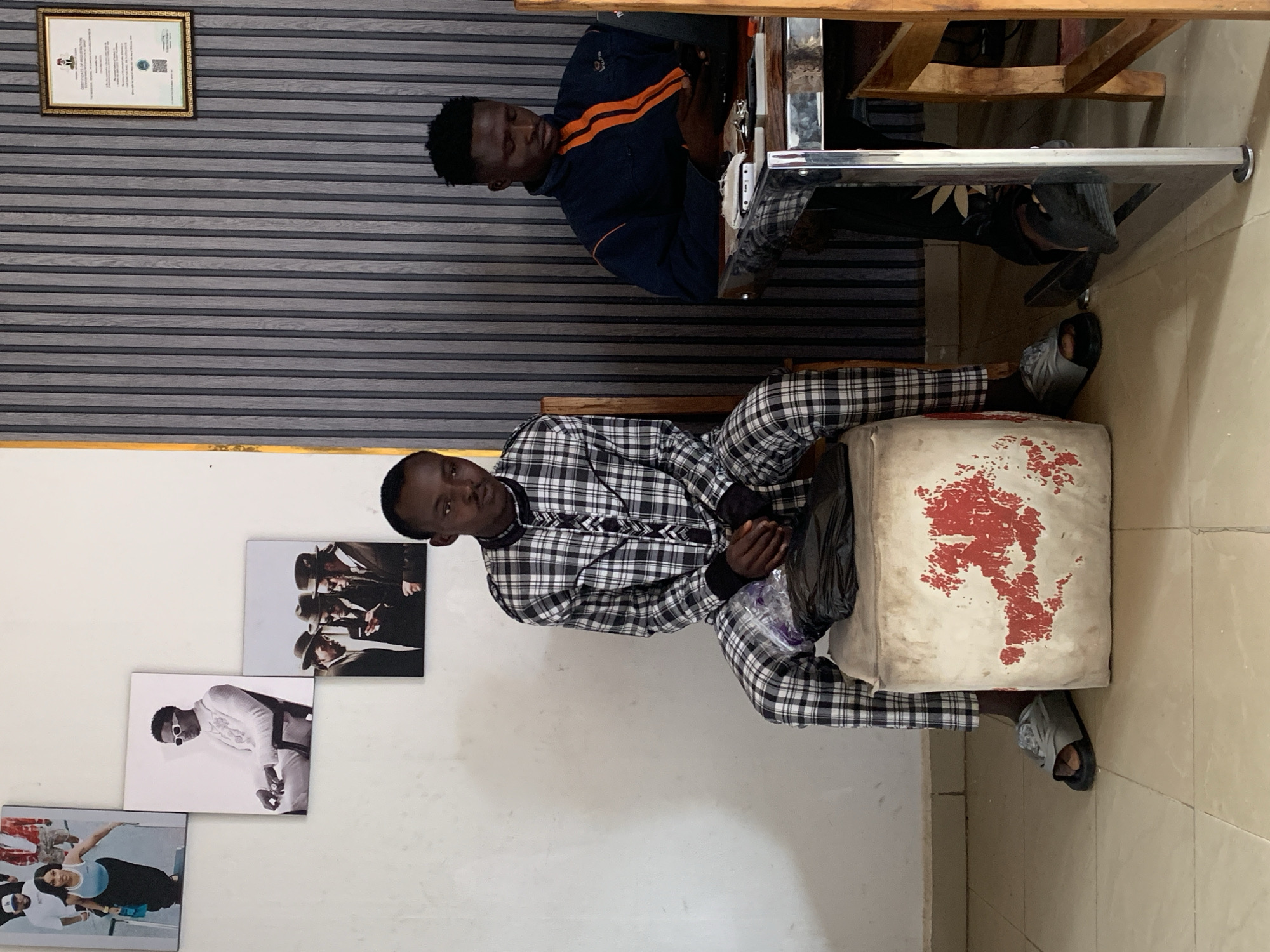 Two young African men are indoors. One man sits at a table with a dark blue jacket that has orange stripes on the shoulder area and sleeves. The other man sits on a beige cushion with red paint stains on it. He wears a gray and white plaid outfit. The wall behind him has three framed pictures arranged vertically. A framed certificate hangs high on the wall behind the first man. The location is in Jos, Nigeria.