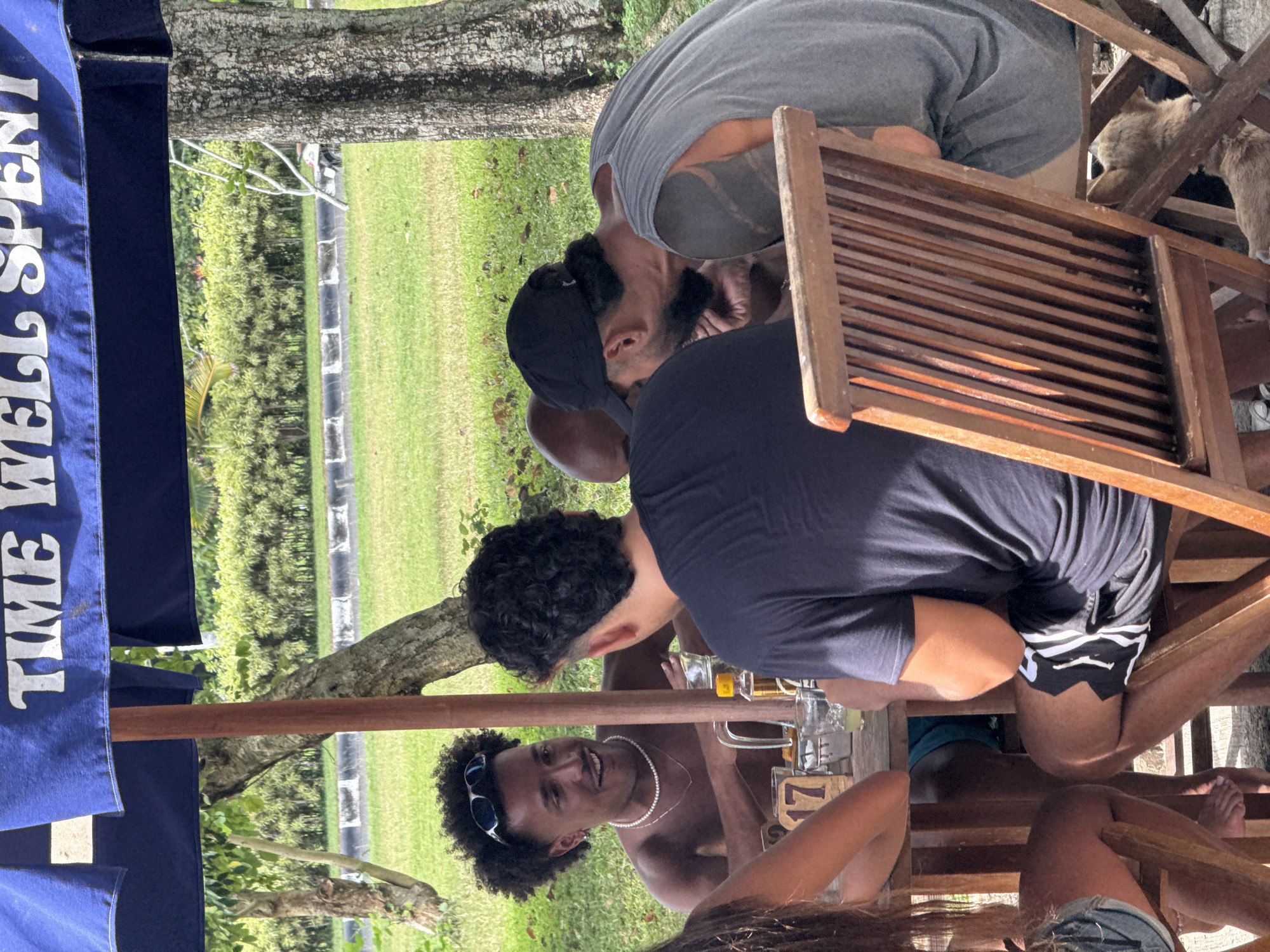 The scene shows a group of people sitting around a table at what appears to be an outdoor restaurant or bar. A blue umbrella with white writing that says "TIME WELL SPENT" is partially visible in the upper left.
The people at the table are in casual clothing, including shorts, t-shirts and a necklace. They are drinking from glasses and appear to be socializing. There are beer bottles and glasses on the table. Some of the people are sitting on wooden chairs. A dog is also visible.
The environment is grassy with trees. The location is Kuta Selatan, Indonesia.