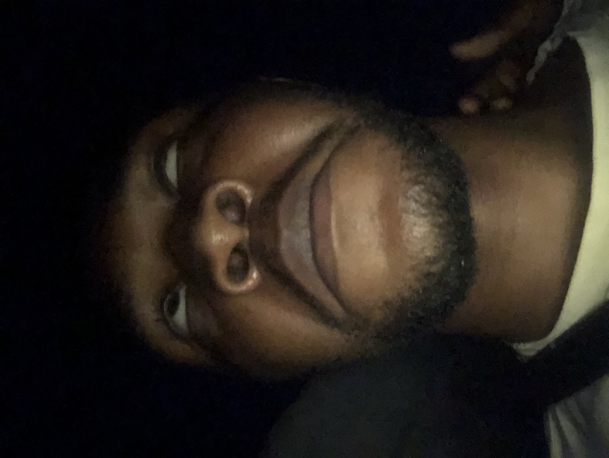 A close-up selfie of a Black man with a dark beard, looking up towards the camera. The background is dark. The location is Unknown, Unknown.