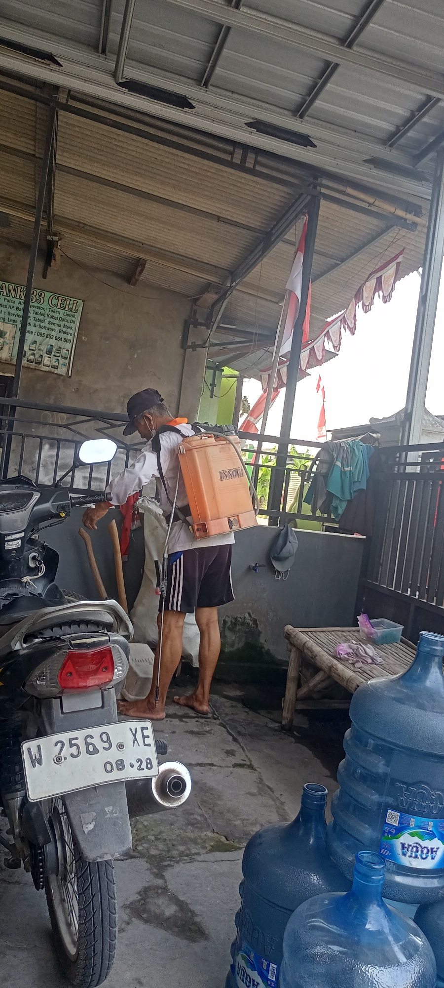 In Gempolklutuk, Indonesia, a man, appearing to be of middle age with weathered skin, stands in a covered outdoor area, possibly a workshop or carport. He is wearing a white long-sleeved shirt, dark shorts with white stripes, a dark baseball cap, and a face mask. Strapped to his back is an orange backpack sprayer with the brand "INNOVA" visible. He holds a long wand, suggesting he is preparing to spray or has just finished spraying.
To his left, a motorcycle is parked, its front tire and license plate, bearing the inscription "W 2569 XE 08.28," are visible. Behind the man, leaning against a sack, are two wooden poles, one with a shovel head. The environment is rustic, with a corrugated metal roof supported by metal beams overhead. A gray wall serves as a backdrop, adorned with a sign for "BANK 33 CELL" advertising various services. Indonesian flags and pennants are hung across the ceiling, indicating a possible national holiday or celebration.
To the right of the frame, a rustic wooden bench sits against a dark wall. On the bench, a folded pink cloth and a blue plastic container are placed. Several large, blue, 5-gallon water cooler jugs are stacked on the ground in the foreground, one of which displays the "AQUA" logo. The ground is a concrete surface, showing signs of dirt and moisture. The overall scene depicts an everyday activity in a seemingly residential or small business setting in Indonesia.