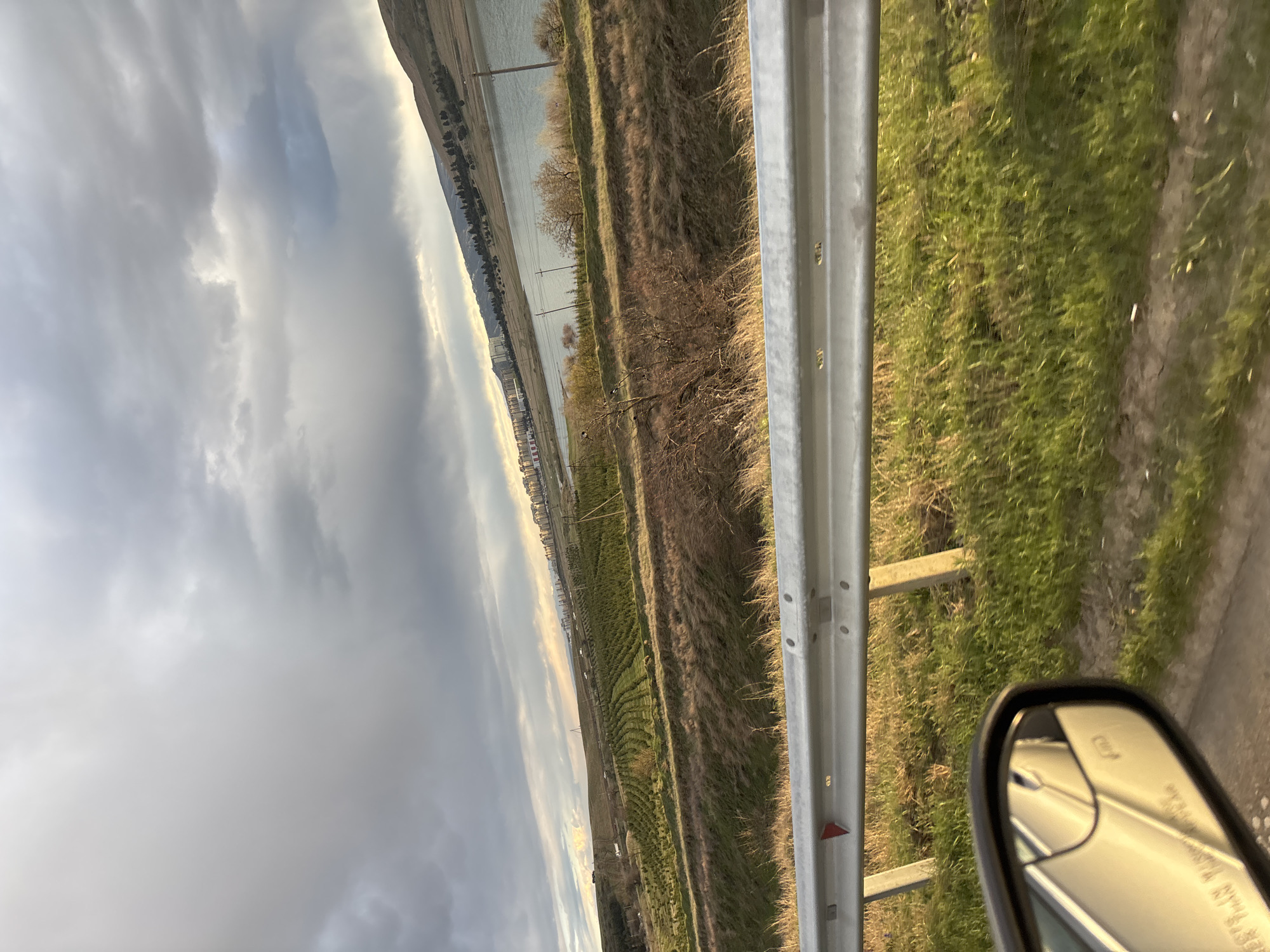 The image depicts a landscape viewed from a vehicle, likely a car, traveling along a road with a metal guardrail. A side mirror is visible in the lower right corner. Beyond the guardrail, there is grassy vegetation and a stretch of water with buildings in the distance, possibly a part of Tbilisi, Georgia. The sky is overcast with heavy clouds.