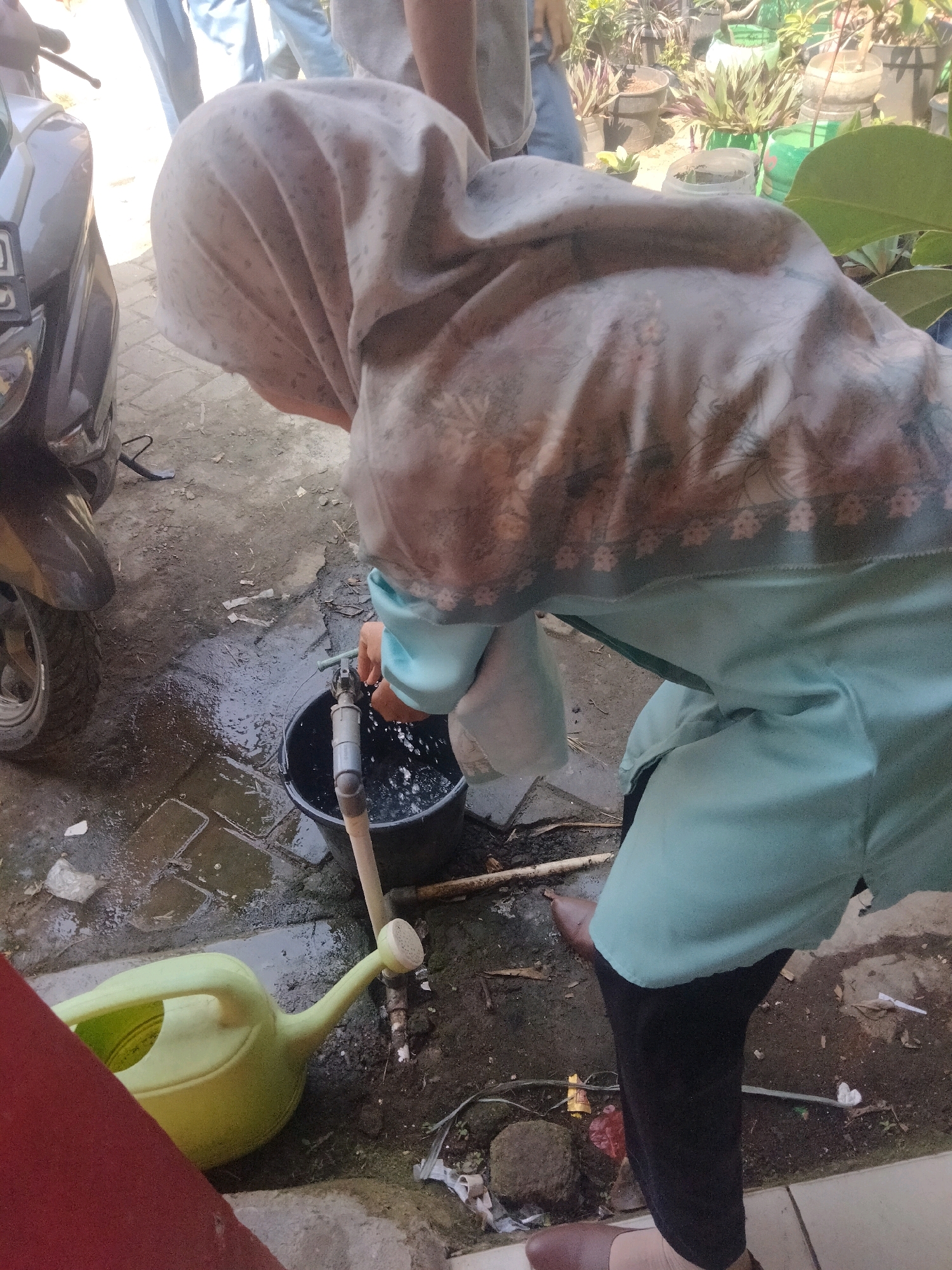 In Suka Sari, Indonesia, a person wearing a light blue hijab and a light blue tunic is filling a black bucket with water from a faucet. Water is splashing out of the bucket and onto the wet ground. Next to the bucket is a light green watering can. A motorcycle is partially visible on the left side of the image, and a variety of potted plants are in the background. The ground is made of a mix of concrete and dirt, and debris is scattered around.