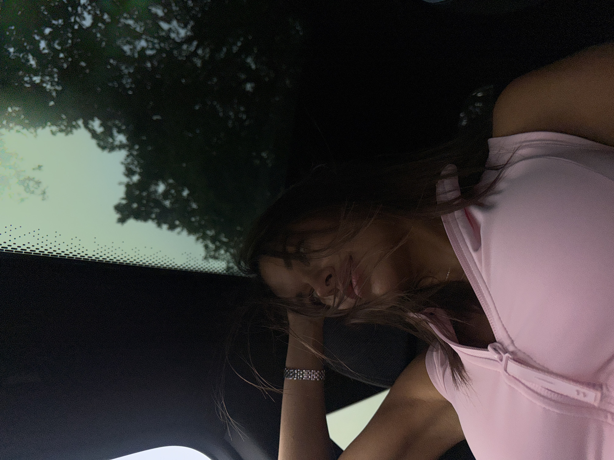 A young woman is seated in a vehicle, looking upwards and to the right. Her right arm is raised, supporting her head. She is wearing a light pink, sleeveless top with a zippered neckline. A silver bracelet is visible on her wrist. Her hair is brown and appears windblown. The vehicle's interior is dark, with a prominent black headliner. Through the windshield, trees and a bright, overcast sky are visible. The image was taken from inside the car, looking up through the windshield. There is no indication of the location being Amsterdam, Netherlands.