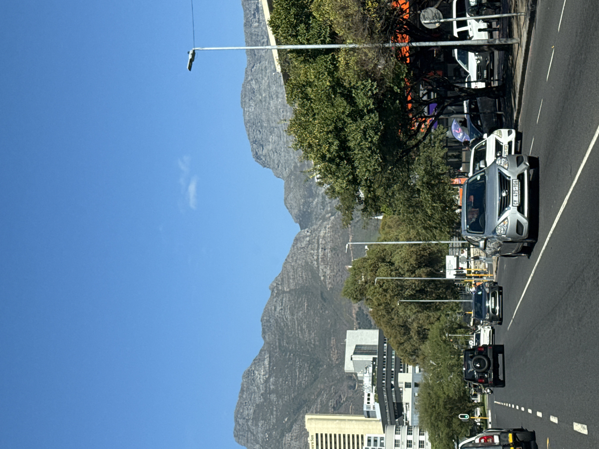 The image captures a street scene in bright daylight, dominated by a majestic, rugged mountain range in the background. The sky is a clear, vibrant blue with a few faint wisps of clouds.
In the mid-ground, at the base of the mountain, are several multi-story buildings, including one light-colored building with the partial text "thern Sun" visible, likely "Southern Sun." Green trees with some orange foliage line the street, partially obscuring the mountain and buildings. Streetlights and traffic lights are also visible.
The foreground and mid-ground feature a multi-lane road with active traffic. A silver SUV, possibly a Nissan Livina, is prominent in the mid-right, displaying the license plate "CY 475-906." Behind it, a white car's blurry license plate begins with "CA 95-864." Further back, a dark sedan or hatchback has a purple banner or flag extending from its passenger window, suggesting a parade, protest, or event. Other vehicles, including a white car and a dark SUV with a rear-mounted spare tire, are visible along the road. A green traffic light signal is visible further down the street. The road features solid and dashed white line markings.
The scene depicts a busy urban environment with a striking natural backdrop, indicative of Cape Town, South Africa, given the "CY" license plate prefix and the prominent mountain resembling Table Mountain. The clear blue sky and bright lighting suggest a sunny day, likely midday or early afternoon.