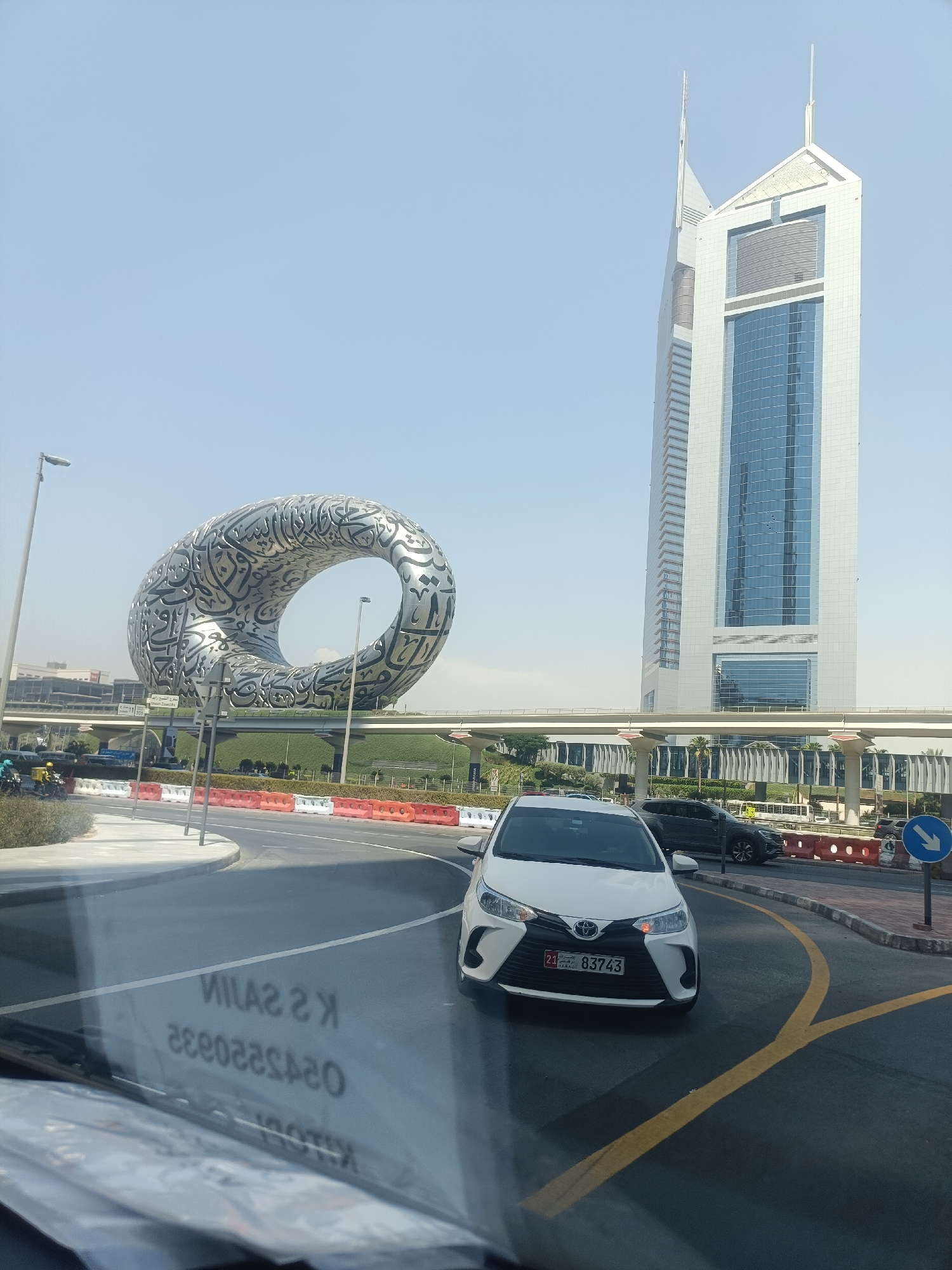 The image depicts the Museum of the Future, a uniquely shaped building with Arabic calligraphy on its facade, in Dubai, United Arab Emirates. Adjacent to it stands the Emirates Tower, a prominent skyscraper. The scene is viewed from a car, with a white Toyota Yaris in the foreground displaying license plate 83743. Traffic barriers and road markings are visible, indicating an urban environment. The sky is clear and blue, suggesting a sunny day. Reflections on the car's windshield reveal text, which appears to be "K S SAIN 0525220832" in a mirrored format.
