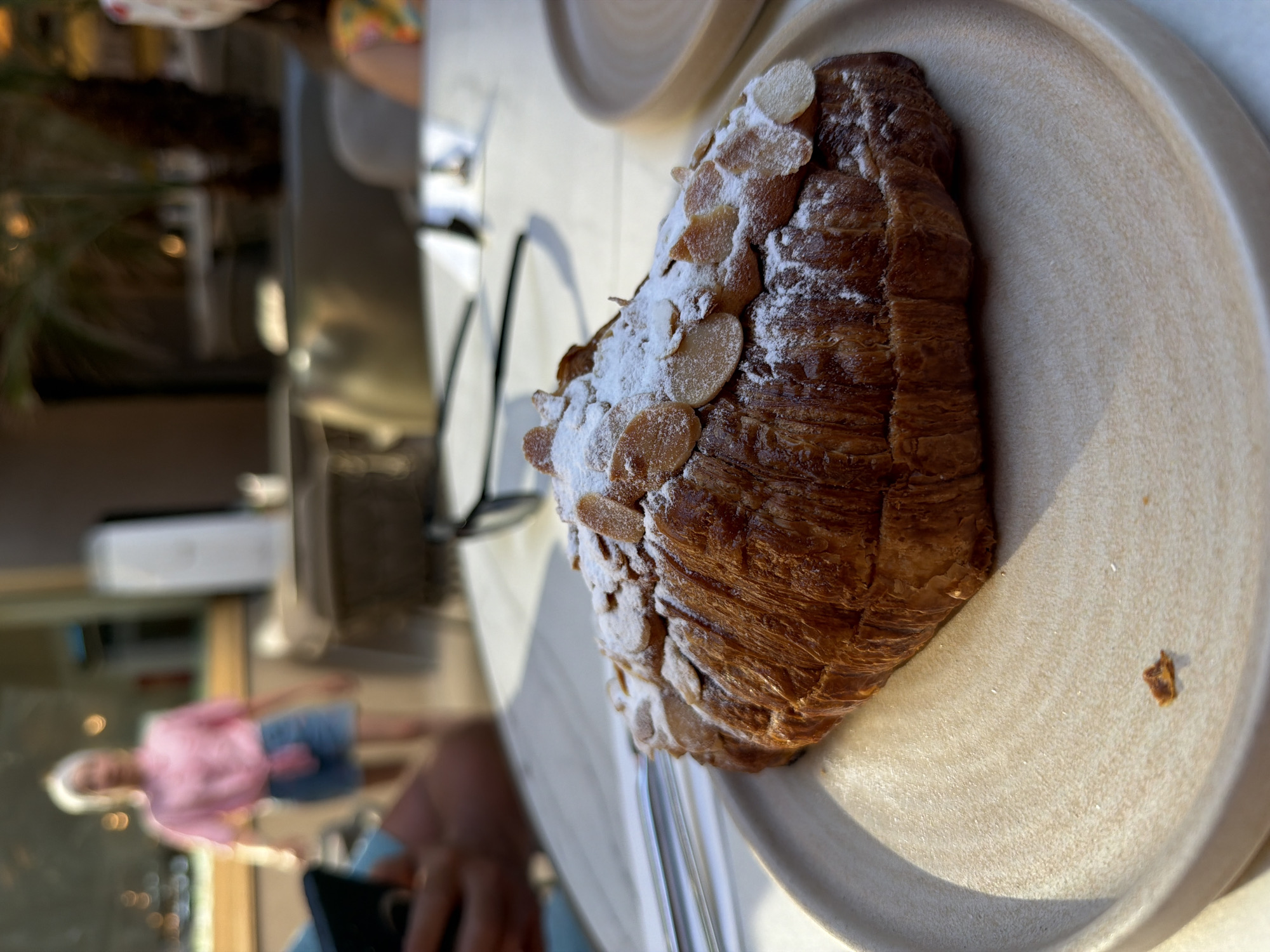A vibrant, close-up photograph centers on a delectable almond croissant presented on an off-white, irregularly shaped ceramic plate. The croissant, golden-brown and flaky, is generously dusted with powdered sugar and topped with numerous slivered almonds, showcasing its rich texture. The plate features a matte, slightly textured surface and a gently raised rim, adding an organic feel to the presentation.
Strong natural light, presumably sunlight, illuminates the scene from the right, casting highlights on the sugary croissant and the plate, while creating soft shadows that enhance depth. The background is intentionally blurred, drawing primary focus to the pastry. Within the out-of-focus background, a person wearing a pink top and denim shorts is visible, suggesting a relaxed social setting. Another blurred hand, holding a black smartphone, is also discernible closer to the foreground.
The setting appears to be a brightly lit cafe or restaurant, possibly outdoors, given the strong natural light. Reflections on a light-colored table and a clear drinking straw lying next to the croissant further contribute to the dining scene. The overall impression is one of a pleasant daytime meal or snack, likely breakfast or brunch, in a casual environment, potentially in Fethiye, Turkey. No text is visible in the image.
