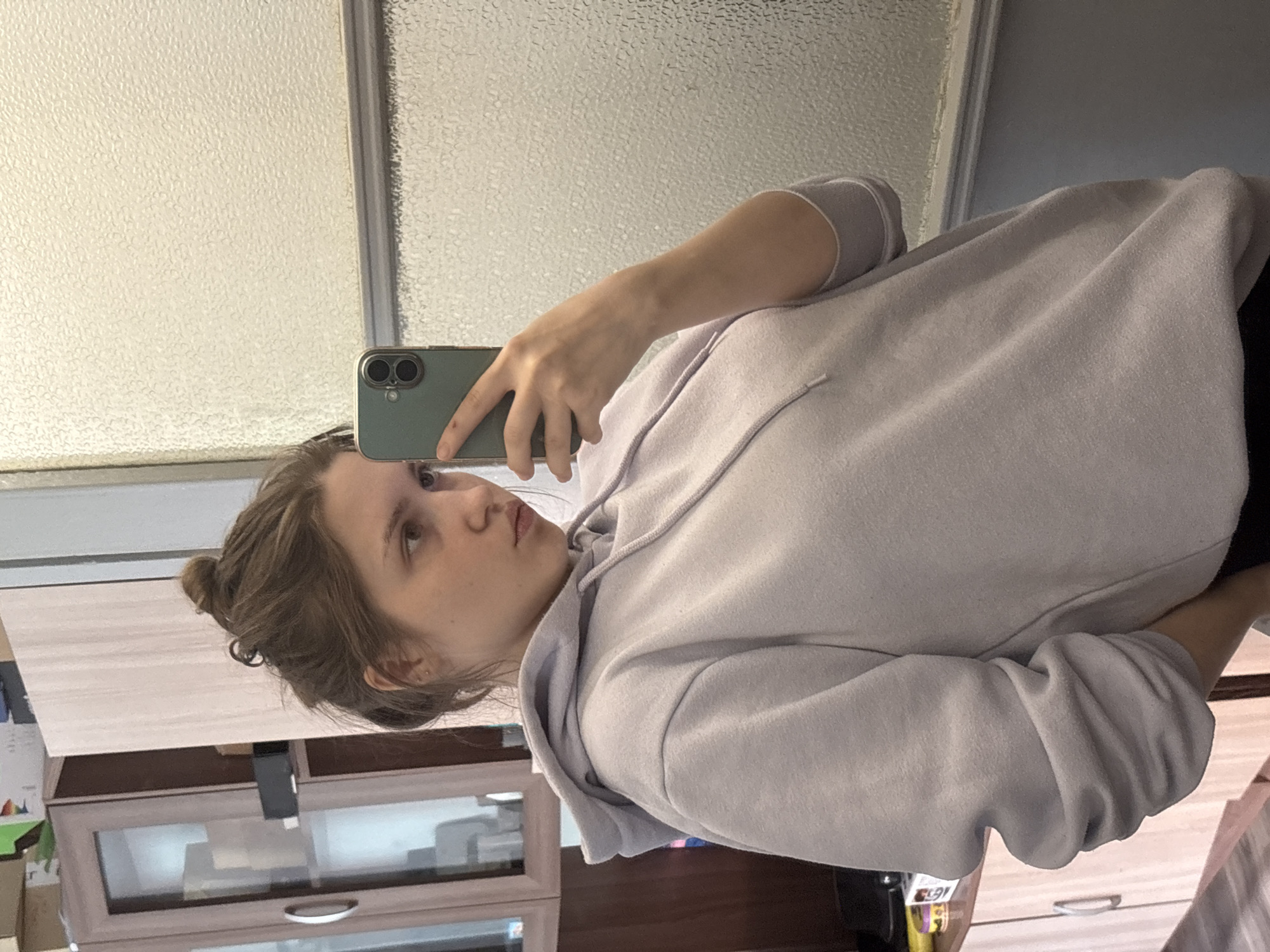 A young woman with her light brown hair in a bun is taking a selfie in what appears to be a bathroom. She is wearing a light purple hoodie and holding a green cell phone. The background shows a frosted glass window and a cabinet with a light wood grain finish. The location cannot be determined to be Saint Petersburg, Russia, from the visual information provided.