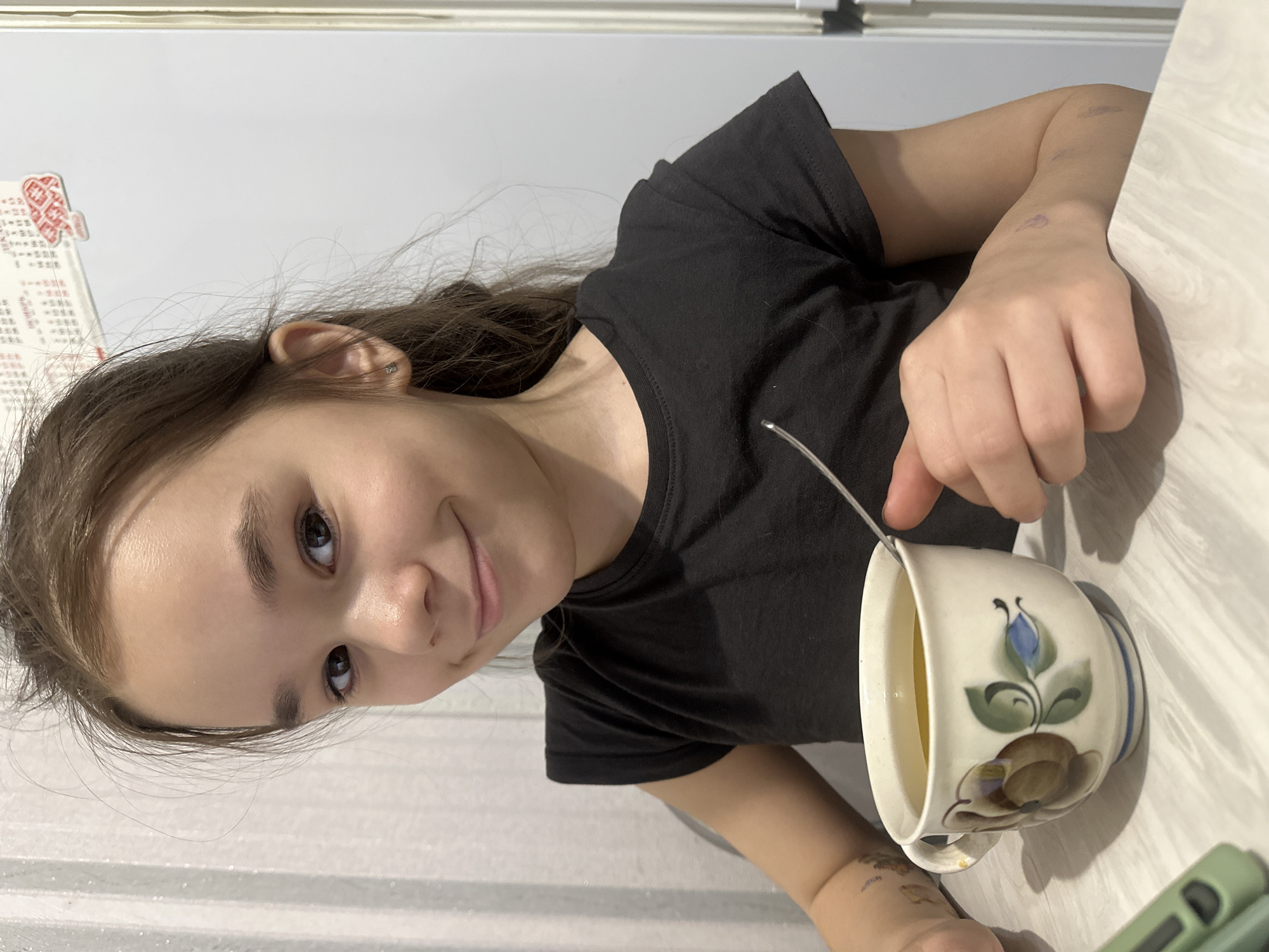 The image, taken in Uva, Russia, captures a young person, likely female, seated indoors at a light-colored table. She has brown hair and is wearing a black t-shirt. She is looking directly at the camera with a subtle, pleasant smile.
Her right hand holds a spoon or stirrer inserted into a large, white ceramic mug. The mug features a hand-painted floral design with a blue flower, green leaves, and brown motifs, along with vertical blue stripes on its side. A light-colored liquid, possibly tea or juice, fills the mug.
Notable details include faint purple marks on the subject's left forearm, which could be pen marks or minor bruises. In the background, a white wall is visible. On the wall, to the upper left of the subject, there's a calendar or poster with red decorative patterns and columns of characters that appear to be Chinese, an interesting visual given the Russian location context. A green phone case is partially visible on the table to the right of the mug. The lighting suggests an indoor setting, and the overall atmosphere is calm.