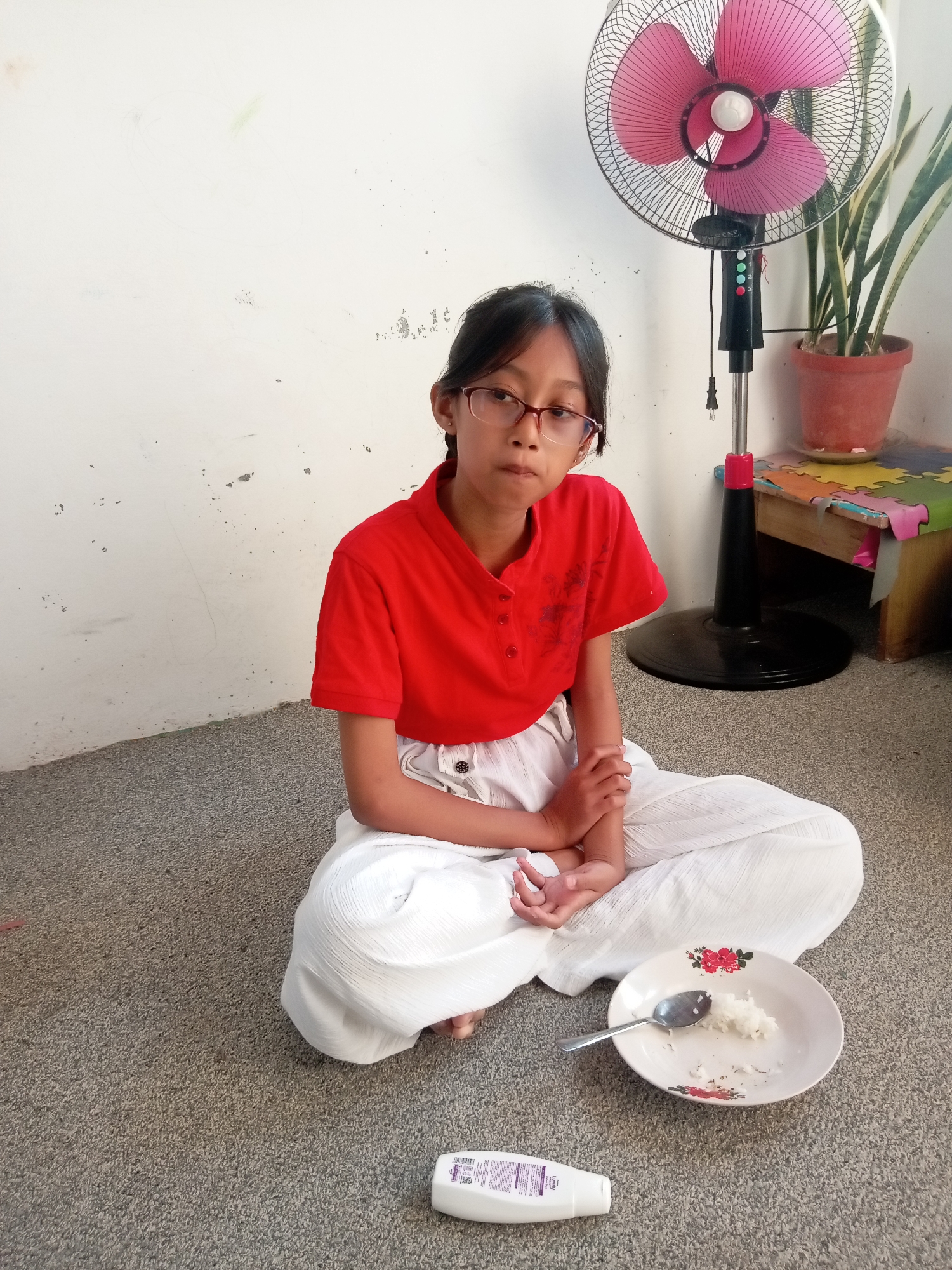 A young girl with glasses is sitting cross-legged on a carpeted floor, looking towards the camera. She is wearing a red t-shirt and white pants. In front of her is a white plate with a small amount of white rice and a spoon. To her right, a standing fan with pink blades is visible, and behind it, a snake plant in a terracotta pot sits on a colorful mat. A white bottle is also on the floor in the foreground. The background is a plain white wall with some scuff marks. This scene appears to be indoors, likely in a residential setting within Batam, Indonesia, given the context.