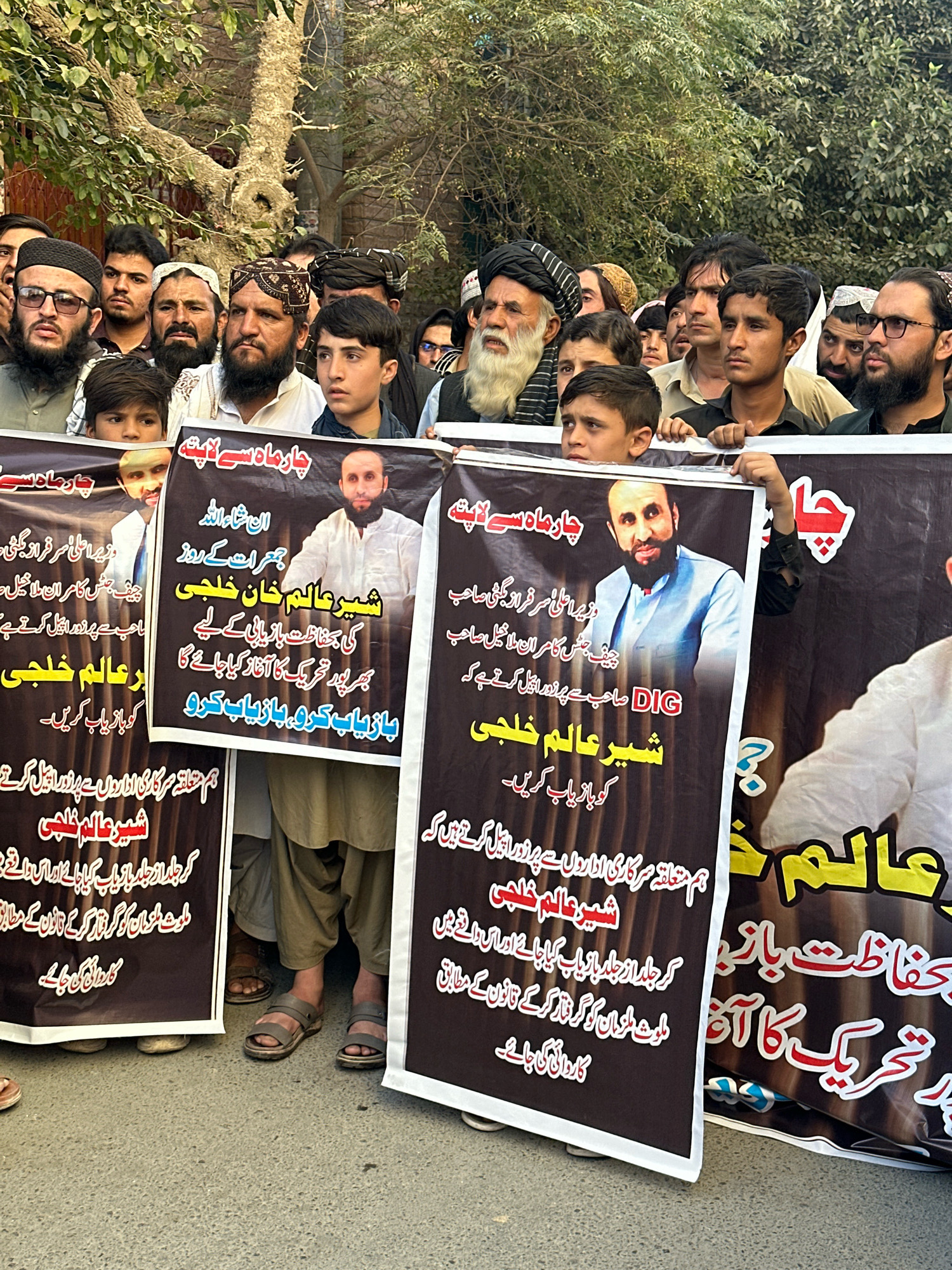 A group of men, some holding banners, are gathered outdoors in what appears to be a street in Quetta City Tehsil, Pakistan. The banners, predominantly black with yellow and white lettering, feature portraits of men and text in Urdu. One banner reads "چهار ماهے" which translates to "four months". Another banner includes the name "شیر عالم خلجی" (Sher Alam Khalji). The individuals are dressed in a mix of traditional Pakistani attire and more casual clothing. Some are wearing head coverings, including turbans and skullcaps. The environment suggests an urban setting with a paved ground and trees in the background. The mood of the gathering seems serious, possibly a protest or demonstration given the banners.