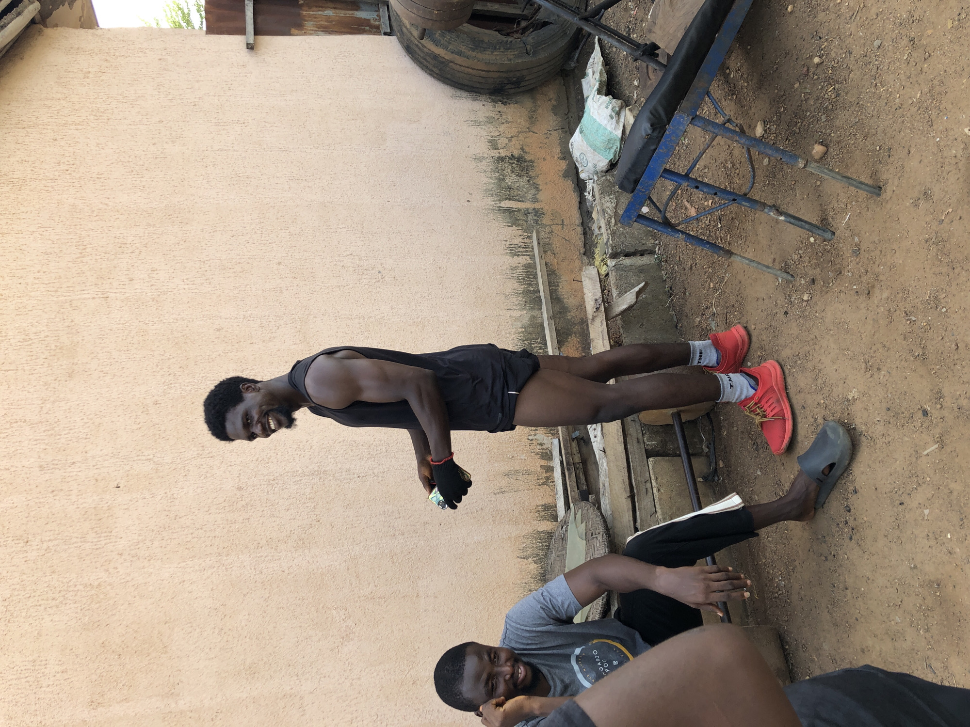 Akwanga, Nigeria: Two men pose outdoors beside a stuccoed wall. One man stands, wearing a black tank top, black shorts, red running shoes with grey socks, and a black weightlifting glove on his left hand. He holds a small object in his gloved hand and smiles. The other man sits on the ground wearing a grey t-shirt and black pants, smiling while looking upwards. In the background are exercise equipment including a tire, weight bench, and a few other metal objects. The ground is dirt with various discarded items.