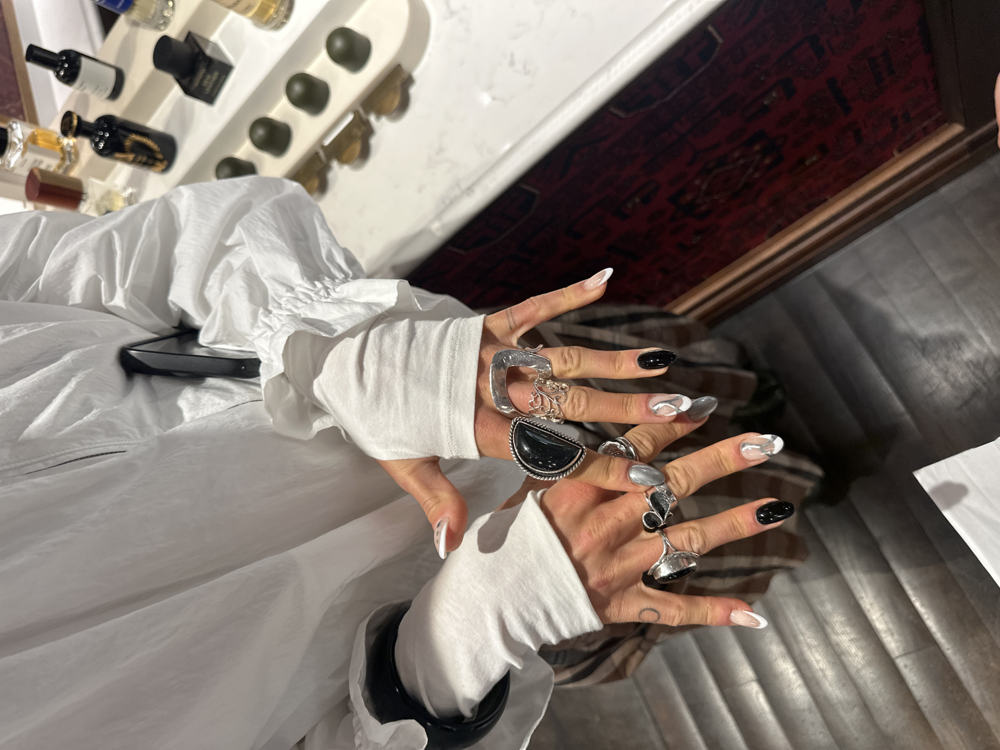 The image shows a person's hands adorned with multiple rings and manicured nails, wearing a white garment with sleeves pushed up to the forearms. A black bracelet is visible on the left wrist. The hands are positioned against a backdrop that includes a surface lined with bottles and spherical objects, a surface with a red patterned design, and a metallic, corrugated surface. A mobile phone can be seen near the left arm of the person. The location in the city of London, United Kingdom is not discernible from the image.