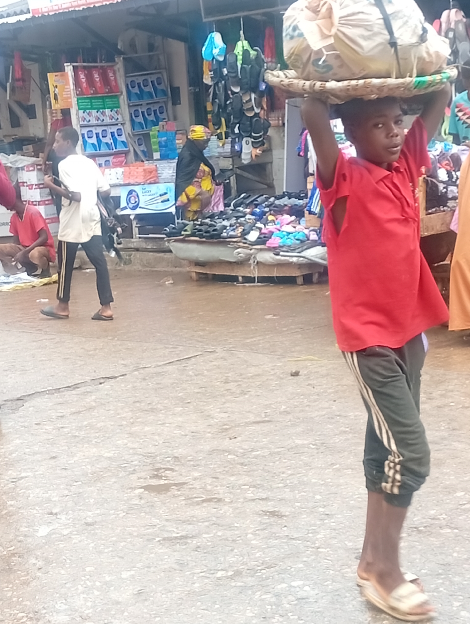 In a street in Maiburiji, Nigeria, a young boy in a red shirt and dark track pants carries a large bundle on his head, balanced on a woven tray. The street is bustling with activity, with people moving around and vendors selling goods. A vendor in yellow head covering stands behind the counter of shoes. A man in a white shirt and dark pants walks away from the boy. Other people are seated or standing nearby. A storefront displays boxes of packaged goods and other merchandise. The ground appears damp or recently cleaned.