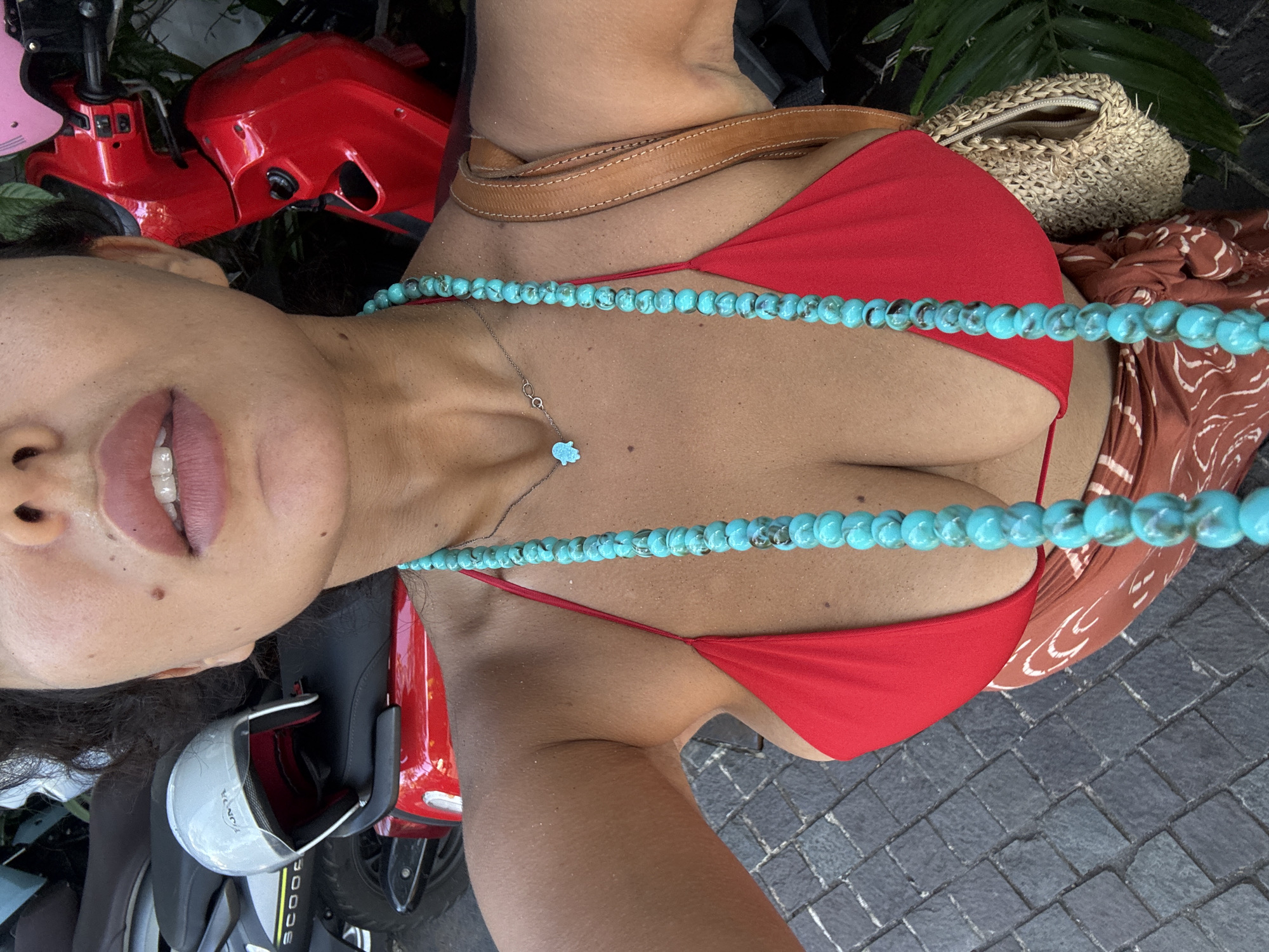 A photograph captures a woman wearing a red bikini and a necklace of turquoise beads, oriented vertically in the frame. The background includes a red scooter, a handbag made of woven straw, and a patterned brown scarf. The location is North Kuta, Indonesia, as indicated by the scooter model, commonly found in that area, and the local Indonesian currency visible in the bag.