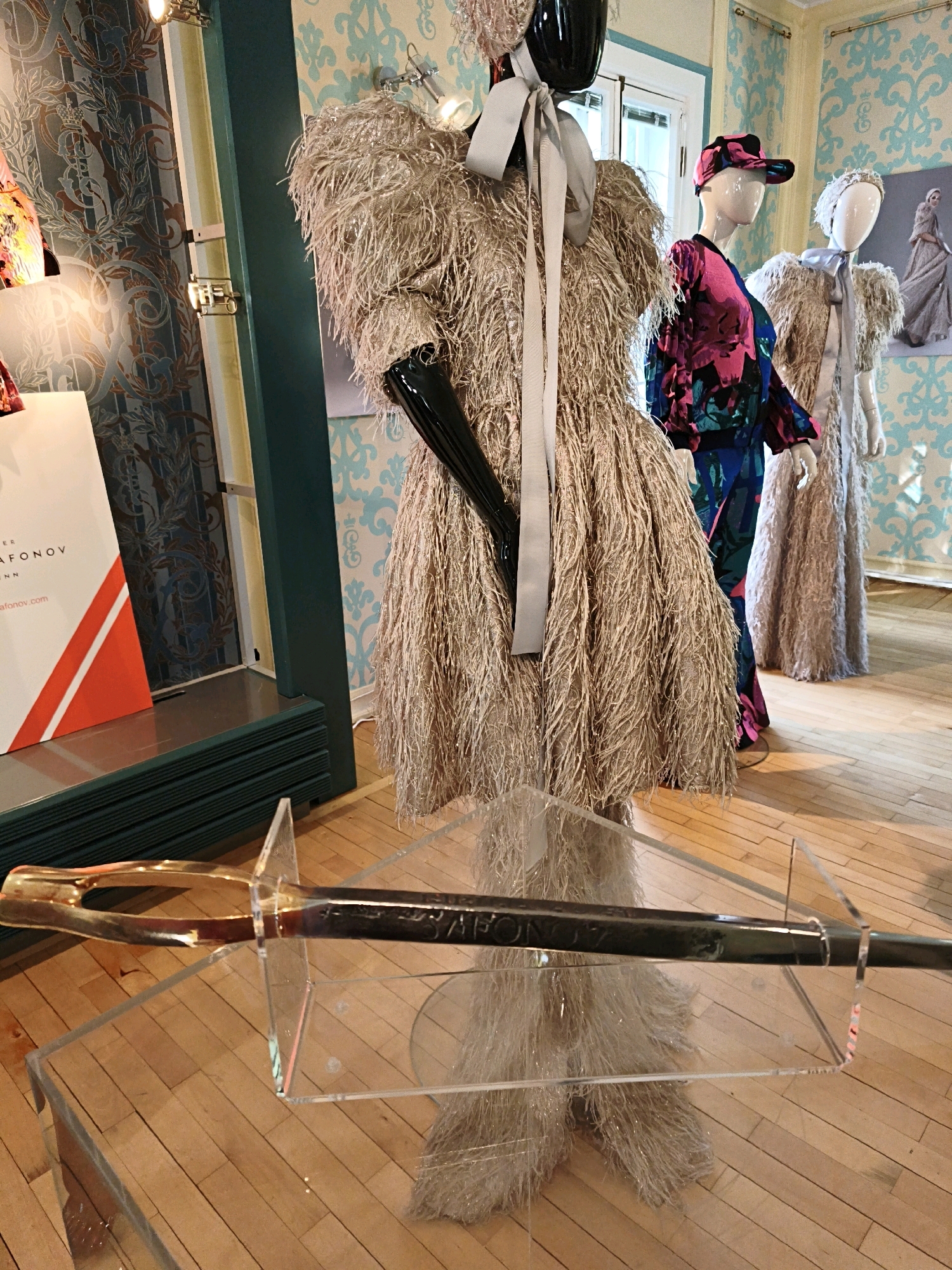 The image depicts a retail display of clothing on mannequins. In the foreground, a sword is displayed on a clear acrylic stand, positioned on a wooden floor. The sword has a gold-colored handle and a dark blade with visible inscription. Behind the sword, three mannequins are arranged. The central mannequin is adorned with a beige, feathery dress and black gloves, with a gray ribbon around its neck. To its right, another mannequin wears a colorful, patterned dress and a pink and black baseball cap. Further to the right, a third mannequin is dressed in a beige, feathery outfit and a light blue ribbon. The background features wallpaper with a blue damask pattern. A white sign with orange accents is visible to the left of the central mannequin. The lighting appears to be from spotlights. The scene is set indoors. The provided image does not contain any information about the city of Tallinn, Estonia.