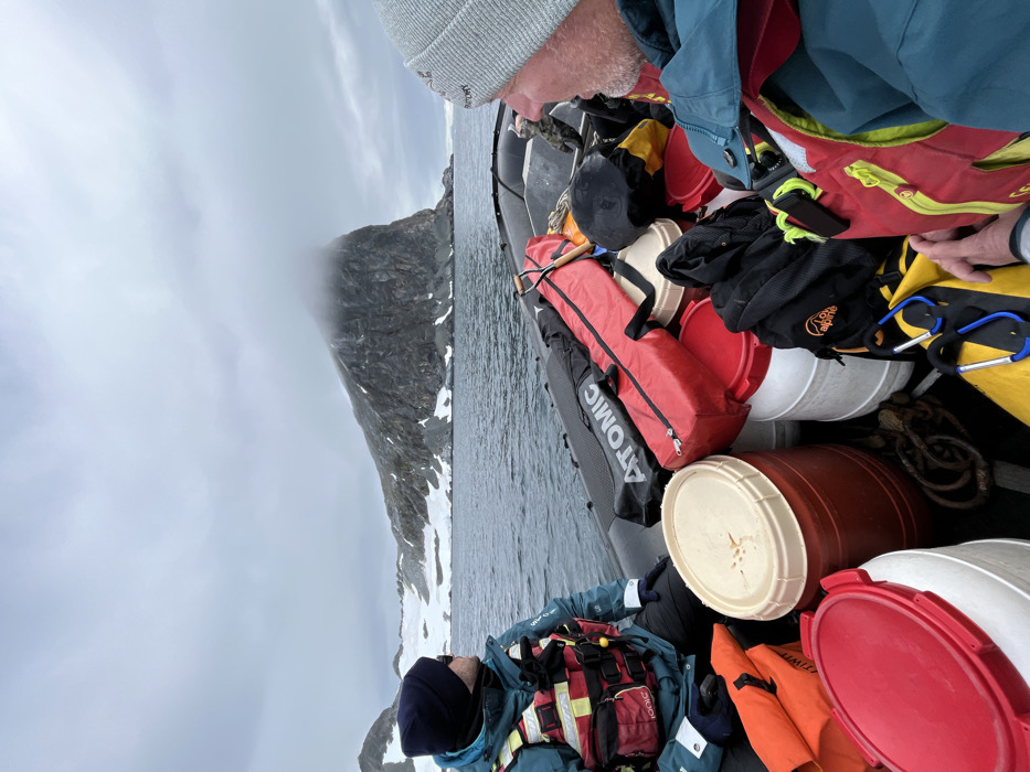 This image captures a boat expedition through a mountainous, cold environment under an overcast sky. The scene is viewed from slightly above and behind the front of a rigid inflatable boat (RIB) or Zodiac, which is heavily loaded with gear.
Two individuals are visible in the boat. One person, positioned closer to the viewer, wears a grey knitted beanie, a blue jacket, and a red life vest, looking towards the distant mountains. Faint text, possibly "ARCTERYX", is visible on their beanie. Further along in the boat, a second person wears a dark blue beanie, a blue technical jacket, and a red and yellow life vest, also facing forward.
The boat is packed with various items, indicating an expedition or transport of supplies. These include several dry barrels or buckets in red, white, cream, and brown colors. A long red bag with "ATOMIC" printed on its side is prominent, as is a black bag bearing the "Lowe Alpine" logo. Ropes, carabiners, and other pieces of yellow and black gear are also stowed in the boat.
The surrounding landscape features dramatic, dark rocky mountains partially covered with snow, stretching along the calm water. Low-hanging clouds or mist obscure the upper portions of the mountains, contributing to a muted, cold, and dramatic atmosphere. The light is diffused, suggesting an overcast day in a high-latitude or alpine region. The heavy clothing and specialized gear reinforce the impression of a journey through a rugged and chilly environment.