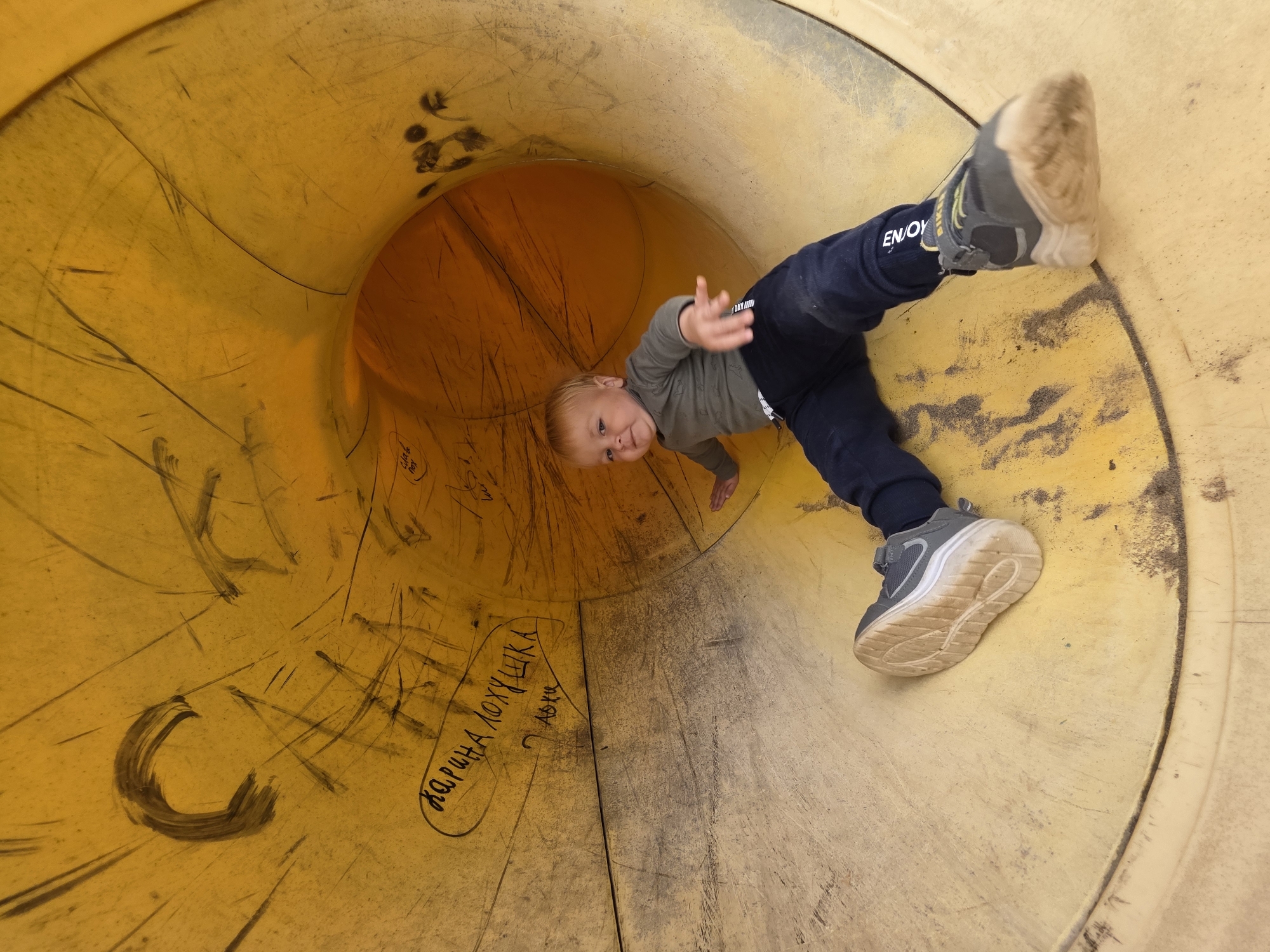 A young Caucasian child with blond hair is sitting inside a bright yellow, tubular playground slide. The child is wearing a long-sleeved grey shirt, dark blue pants, and grey sneakers. The slide is made of plastic and shows signs of wear, including scratches and black graffiti. The graffiti includes words written in Cyrillic script, such as "КАРИНА ЛОХУШКА" which translates to "Karina the sucker". The child is looking towards the camera with a neutral expression and is positioned on their right side, with their left leg extended further into the slide. The background is the interior of the slide, which appears to be made of a textured yellow material. The lighting suggests an outdoor setting, likely a playground. The scene appears to be located in the city of Chaikovsky, Russia, given the presence of Cyrillic graffiti.