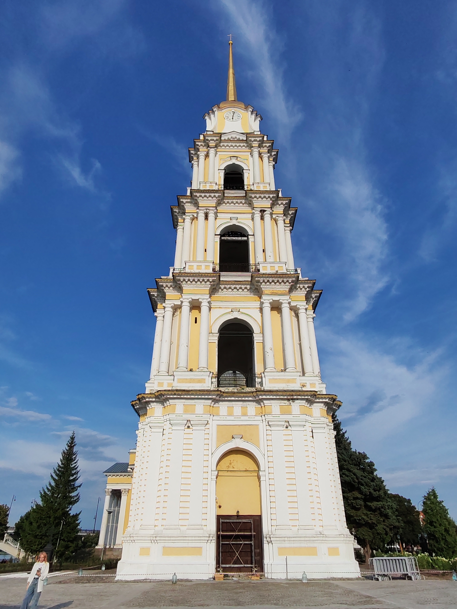 A low-angle, full shot captures a towering, ornate yellow and white bell tower against a bright blue sky with wispy white clouds. The tower features multiple levels with arched openings, Corinthian columns, and decorative cornices. A golden spire tops the structure, crowned with a cross. A clock face is visible on the upper section of the tower. In the foreground, a paved area extends around the base of the tower, with a few trees and a glimpse of a building with yellow accents to the left. To the far left, a young woman with blonde hair, wearing a white blazer over a white shirt and blue jeans, stands and poses, looking towards the camera. To the right of the tower, a dense cluster of dark green trees is visible, and further back, a white metal bench or barrier is present. The scene appears to be an outdoor public space in the city of Bol'shie Mkhi, Russia.