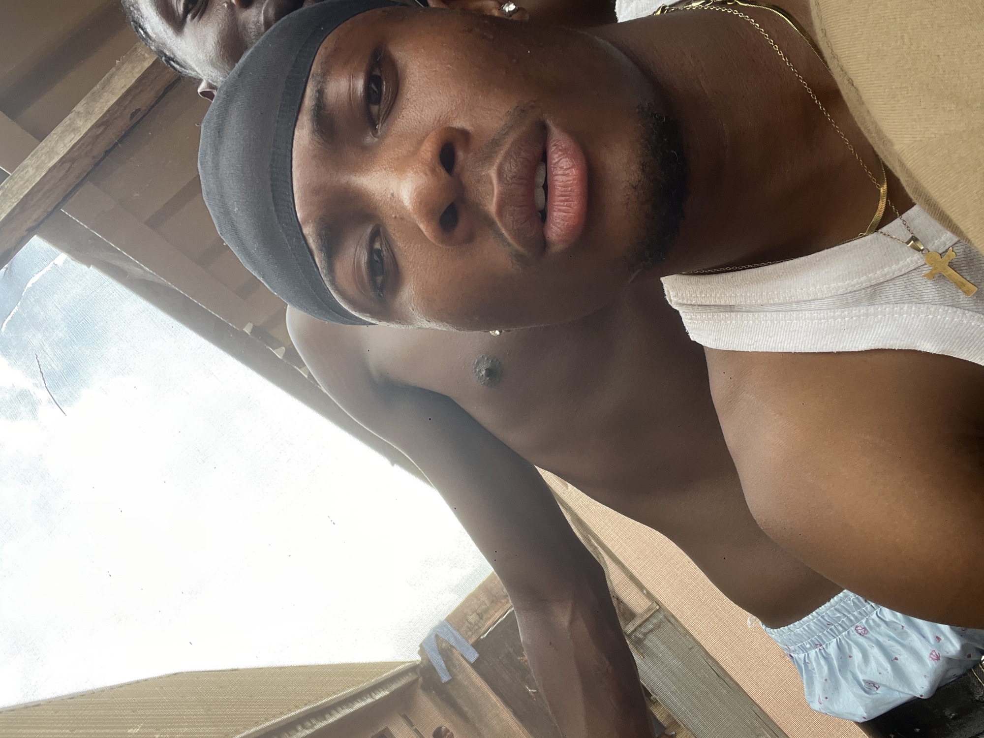 A close-up, low-angle selfie captures two individuals in what appears to be an outdoor, possibly informal setting in Durumi, Nigeria. The foreground is dominated by the face of a young man wearing a black durag and a white tank top. He has dark skin, a closely trimmed mustache and beard, and wears a gold chain with a crucifix pendant and another chain. He has diamond stud earrings. His expression is neutral, and he is looking directly at the camera.
Behind him, another individual, also with dark skin, is partially visible. Their torso is bare, and they appear to be wearing light blue patterned shorts. The background consists of wooden structures and a bright, overcast sky, suggesting an outdoor environment. The overall lighting is bright, with some evidence of wear or dirt on the surfaces. The angle of the shot suggests the selfie was taken from a low position, perhaps from the ground or a lower level of a structure.