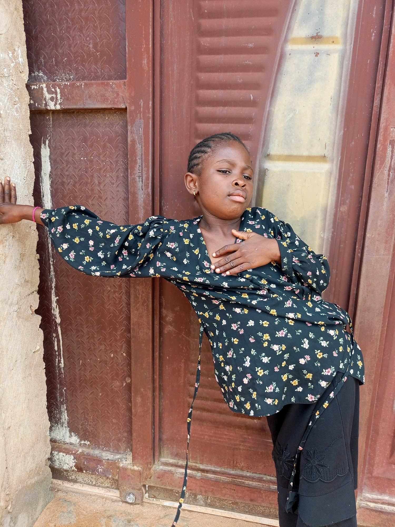 In Tempure, Nigeria, a young girl with braided hair, wearing a black floral top and black pants, poses with her left hand resting on a rough, light-colored wall. Her right hand is placed on her chest. She stands in front of a weathered, reddish-brown metal door. The ground is a mix of dirt and sand. The scene appears to be outdoors, possibly in an alleyway or near a building entrance.