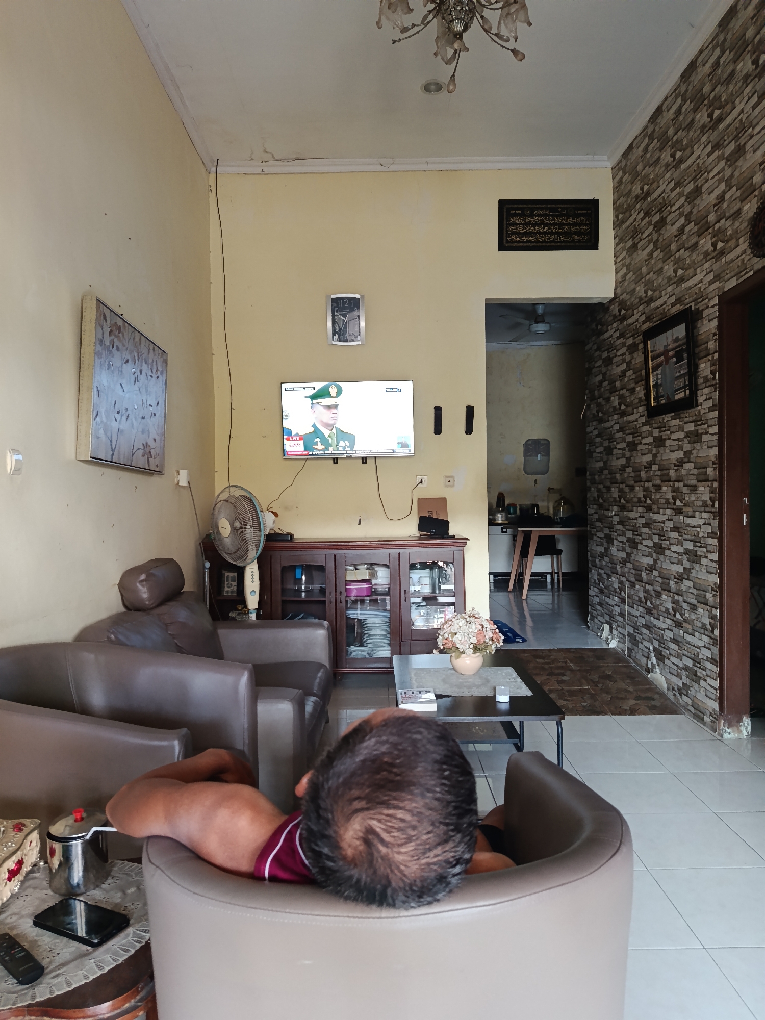 The image depicts an interior scene of a living room in Keboananom, Indonesia. A man is seen reclining on a tan leather armchair, facing away from the camera and towards a television mounted on the wall. The television displays a news program featuring a man in a military uniform wearing a green cap. To the left of the television, a framed artwork hangs on a pale yellow wall, and a white wall-mounted light fixture is also visible. Below the television, a dark brown wooden cabinet with glass doors displays various items, including dishes and decorative objects. A standing fan is positioned to the left of the cabinet.
In the foreground, a dark coffee table is placed in front of the armchair. On the table, a vase filled with artificial flowers, a book, and a small white glass are present. A smartphone and a remote control are visible on a smaller side table to the left, which also holds a metal pot with a red lid.
The floor is covered with light-colored square tiles. To the right of the living room area, a stone-tiled wall extends towards a doorway. Beyond the doorway, a glimpse of a kitchen or dining area can be seen, with a table and chairs. A framed Islamic calligraphy artwork is mounted on the yellow wall above the doorway leading to this area. The ceiling is white and shows signs of minor water damage in some areas. The overall ambiance suggests a casual domestic setting.