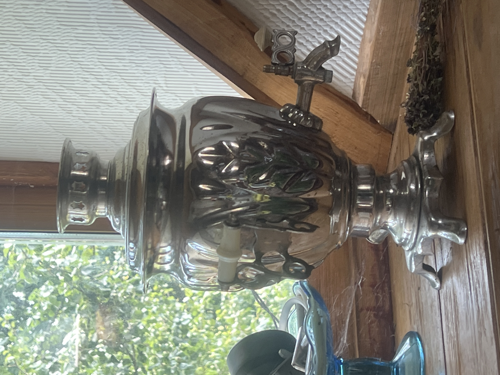 This image depicts a vintage samovar, a traditional metal container used to heat and boil water for tea, placed on a wooden surface. The samovar is ornate, with decorative engravings of leaves and floral motifs, and features a spigot for dispensing water. It is positioned near a window, with lush green foliage visible through the glass, suggesting an outdoor setting. To the left of the samovar, a blue glass dish holds some objects. To the right, a bundle of dried herbs rests on the wooden surface. The background consists of a white textured wall and wooden beams, indicating an interior space. The overall scene evokes a sense of rustic charm and historical ambiance, likely within a home or a heritage site in Norviliškės, Lithuania.