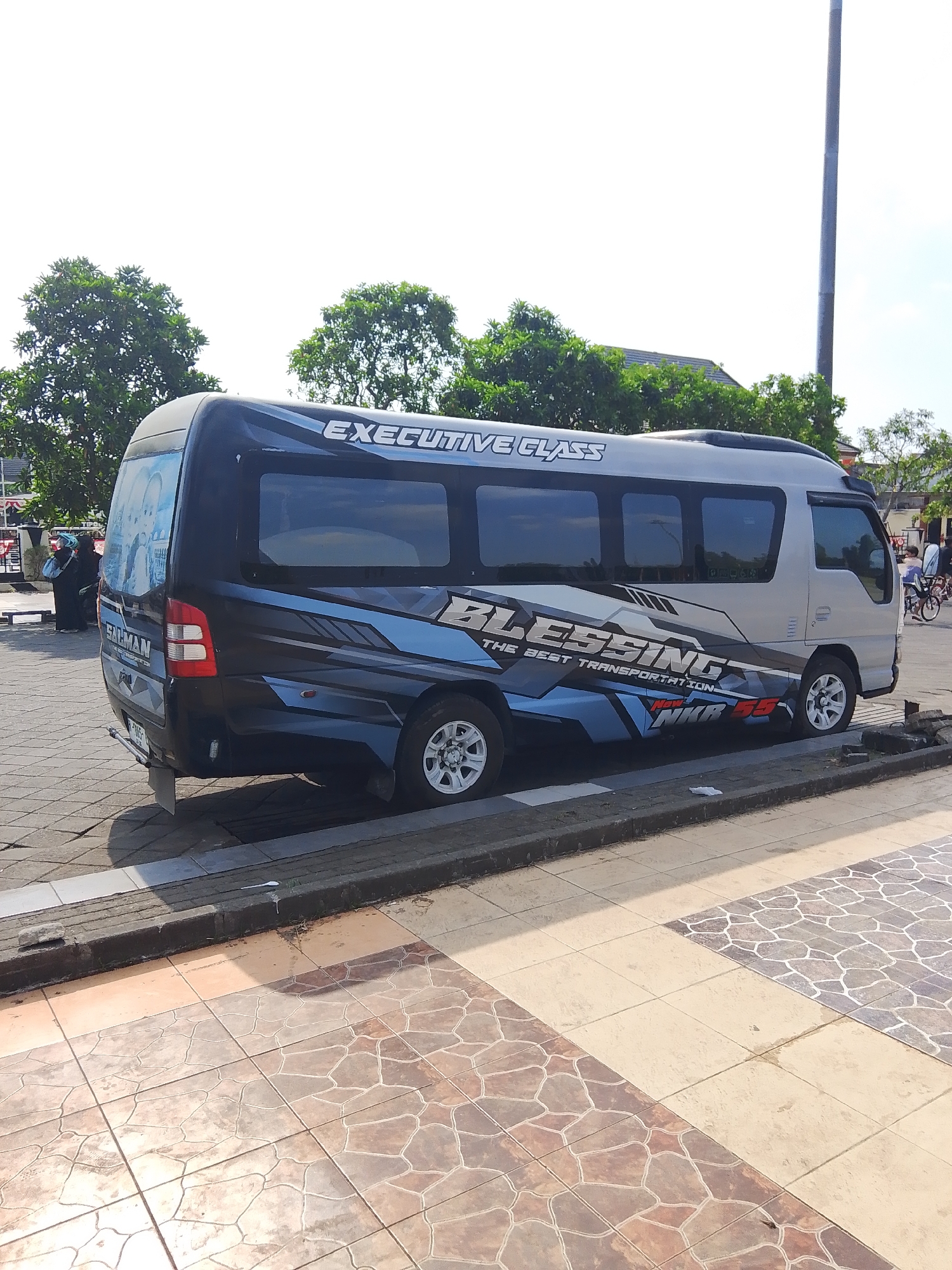 The image depicts a silver and black minibus with the word "BLESSING" and "EXECUTIVE CLASS" written on its side, parked on a paved area in Demak, Indonesia. The bus has tinted windows and a graphic design of blue and black lines. The license plate is partially visible. In the background, there are trees, a tall pole, and some buildings. A few people are visible in the distance, including two individuals in black clothing near a bench. The ground is paved with patterned tiles and there is a curb in front of the bus. The sky is clear and bright, suggesting it is daytime.