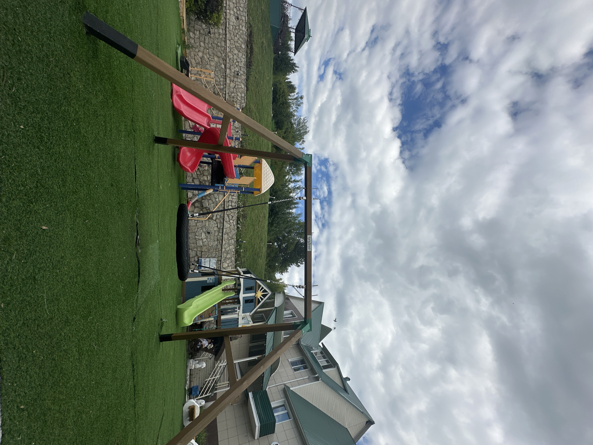 The image depicts an outdoor playground with a swing set, slides, and a playhouse. The ground is covered in artificial green turf. In the background, there is a stone wall, a grassy hill, trees, and a large building with a green roof. The sky is filled with white and gray clouds. The scene is located in Даниловка (алтайский), Russia. There are no people visible in the image.