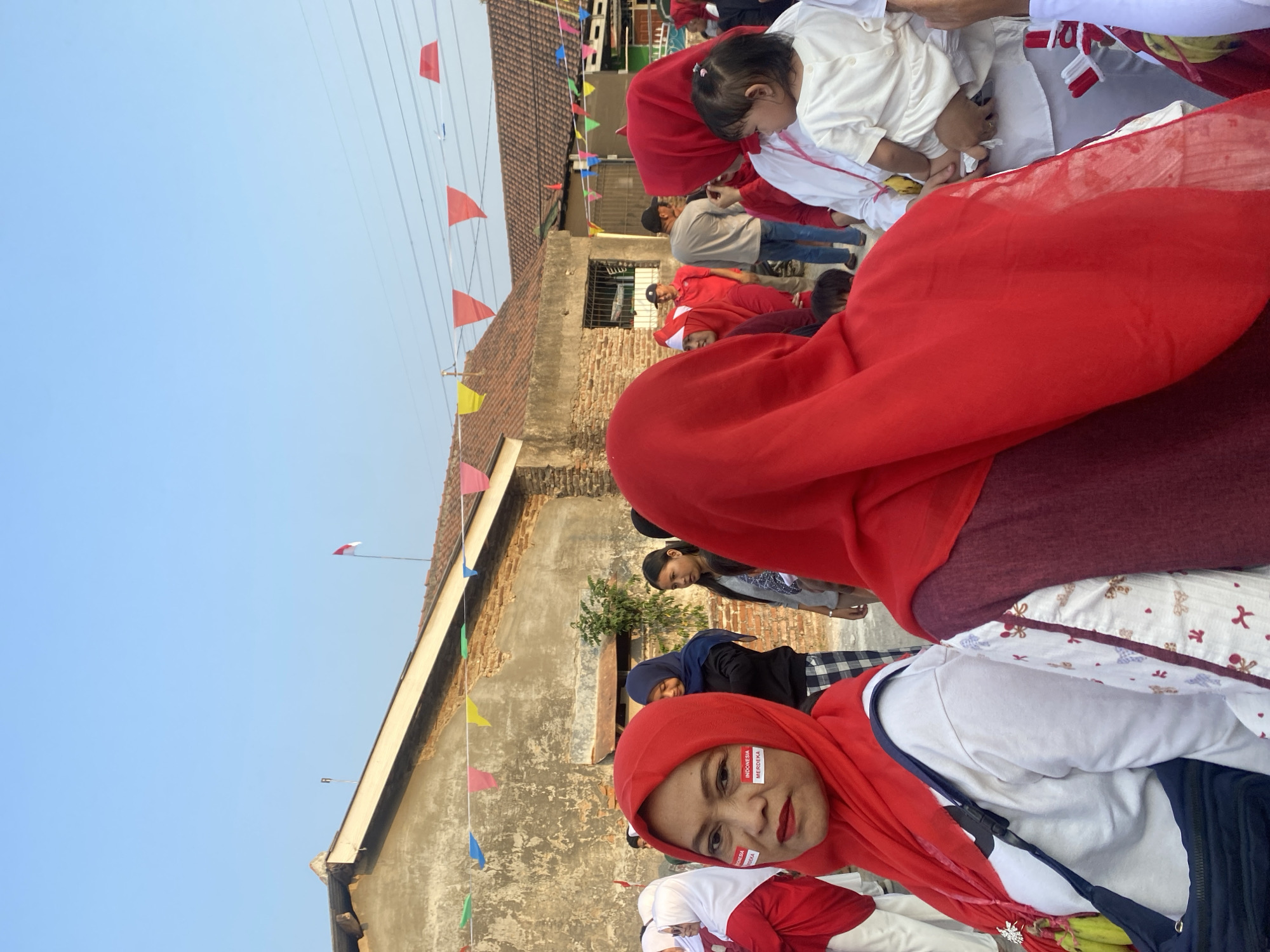 In Cilegon, Indonesia, a woman wearing a red hijab and white clothing is in focus, with face paint on her cheek that reads "INDONESIA MERDEKA". She is part of a crowd gathered outdoors, with a brick building in the background. Many people in the crowd are also wearing red and white, suggesting a celebration or national event. Stringed flags, predominantly red and white, are visible in the background, further indicating a festive or patriotic occasion. The scene appears to be a street or communal area, with a clear sky above.