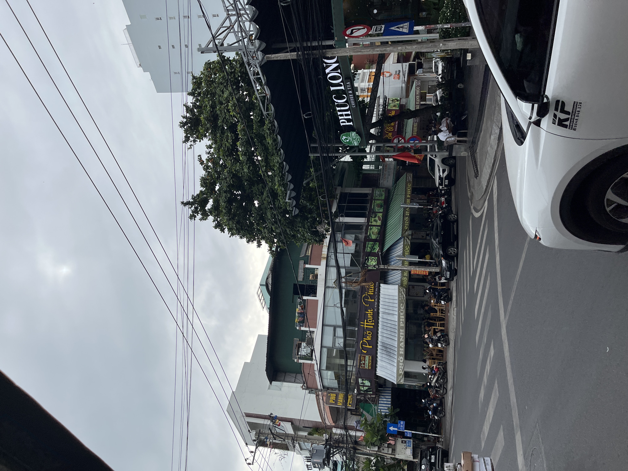 The image depicts a street scene in Nha Trang, Vietnam, likely on a cloudy day given the overcast sky. The foreground shows a white SUV with a black logo on its side. The middle ground features a street with a pedestrian crossing, several motorcycles parked, and a few people seated at outdoor tables in front of a restaurant labeled "PHO HANH PHUC." Further back, there are multi-story buildings housing various businesses, including another "PHO HANH PHUC" and a "PHUC LONG COFFEE & TEA." Overhead, a dense network of electrical wires crisscrosses the sky, a common sight in Vietnamese cities. A large tree with lush green foliage is visible on the right side of the frame. The architecture suggests a commercial area, typical of urban Vietnamese street life.