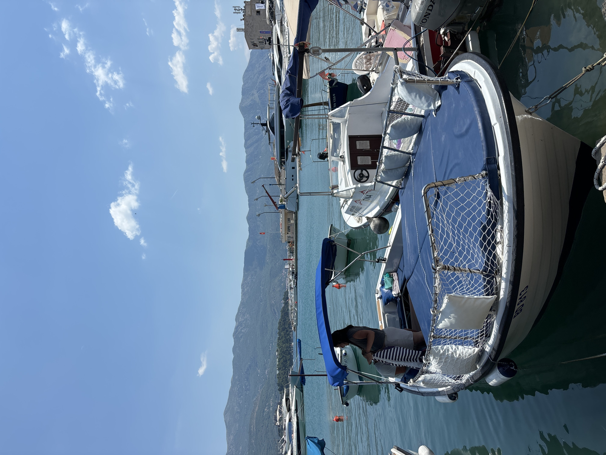A bright, sunny day in Budva, Montenegro, with a clear blue sky dotted with white clouds. The scene is a harbor filled with various boats. In the foreground, a white motorboat with a blue canopy is docked. A woman is standing on this boat, holding a striped towel. To the right, another white boat, named "TINA", is partially visible. In the background, a line of larger yachts and motorboats are moored. Beyond the harbor, a range of mountains forms a scenic backdrop, with some buildings visible on their slopes and at the harbor's edge. The water is a calm, deep blue.