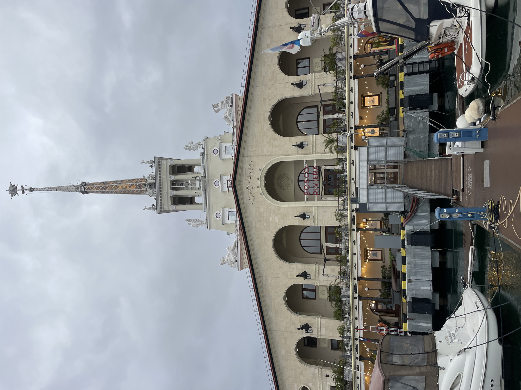 The image depicts the Sochi Sea Port building in Sochi, Russia. The building is a light-colored, multi-story structure with arched windows and a spire. There are several statues on the roof. The building is located next to a marina. Several boats are docked along the marina. The sky is overcast.