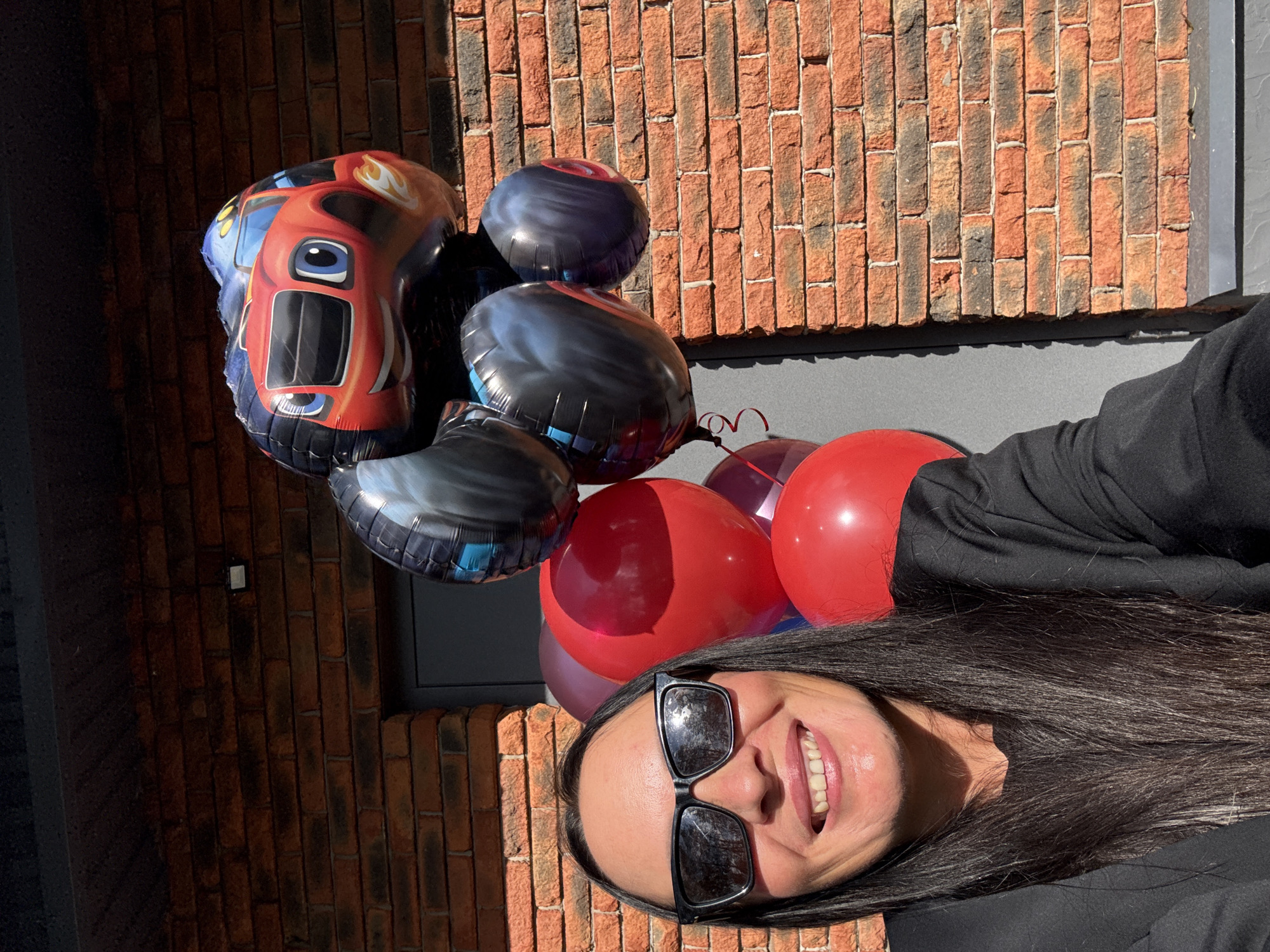 A woman with long dark hair and sunglasses is smiling at the camera. She is wearing a black jacket over a black t-shirt. She is standing in front of a brick wall, and there are several balloons behind her, including one shaped like a red monster truck. The scene appears to be outdoors, possibly in a city. The red brick wall suggests an urban environment. The lighting indicates it is daytime, with sunlight casting shadows. The presence of balloons suggests a celebration or event. The overall atmosphere is cheerful and informal. There is no information in the image to determine the specific location within the city of Chaikovsky, Russia.