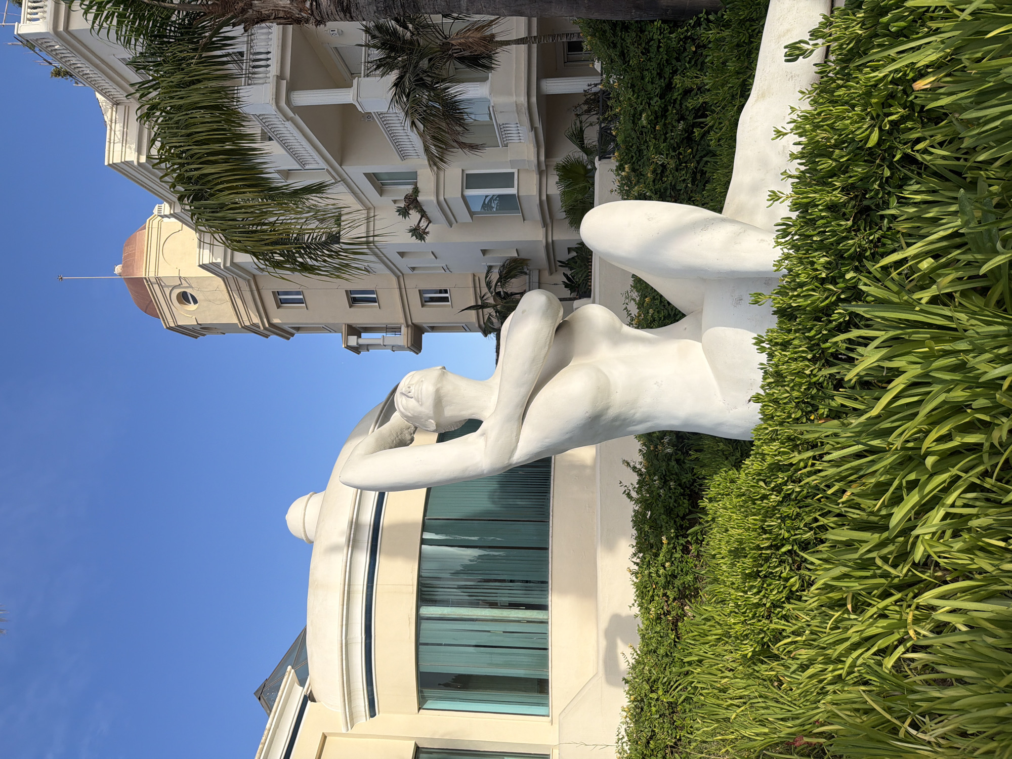 A white, abstract sculpture of a reclining female figure is positioned in a garden. The sculpture is the focal point, depicted with smooth, flowing lines, with one arm raised above its head and the other bent at the elbow. It rests on a bed of lush green foliage, with taller, blade-like plants in the foreground. Behind the sculpture and garden, a pale yellow building with architectural details like balconies, columns, and a domed cupola is visible. A palm tree with vibrant green fronds partially obscures the building. The sky is clear and bright blue. This scene is located in Estepona, Spain, specifically at the entrance to the Luxury Hotel Las Dunas, which is situated in the El Saladillo area of Estepona.