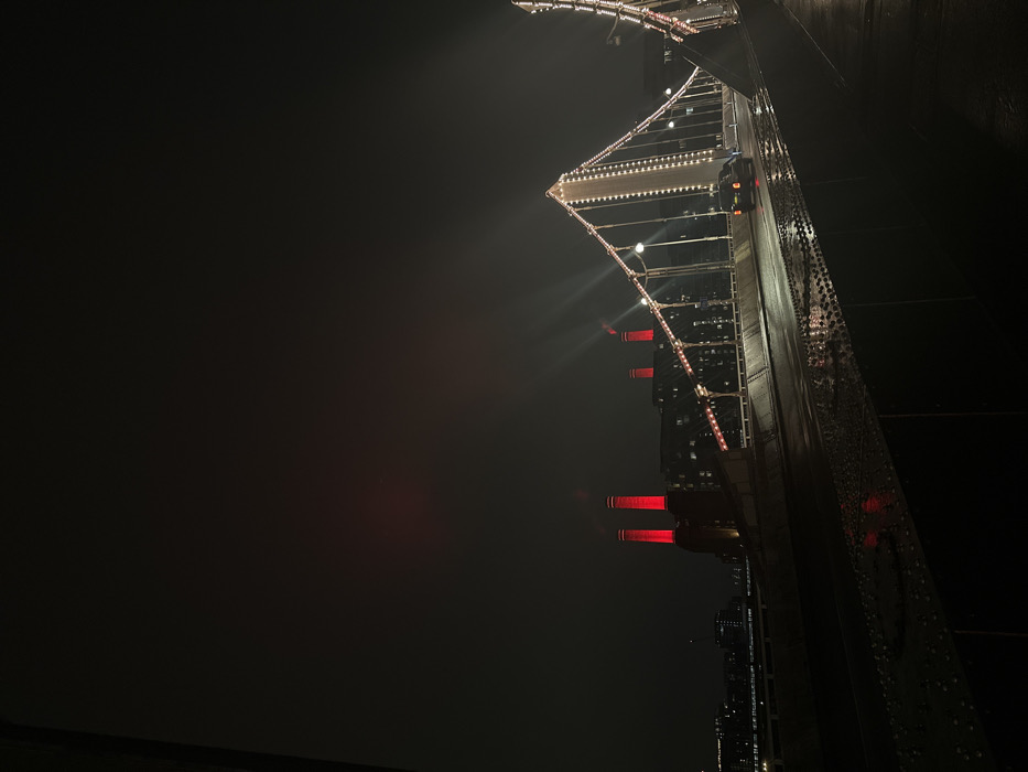 An upward-facing night shot highlights an illuminated bridge or elevated structure. The structure's edge, densely adorned with lights, extends diagonally across the frame, contrasting with the surrounding darkness. The background is predominantly black, suggesting a dark sky or heavy fog. Elements reminiscent of buildings or industrial structures with bright red lights are visible, adding to the eerie atmosphere. The photograph's perspective emphasizes the height and perspective of the illuminated structure.