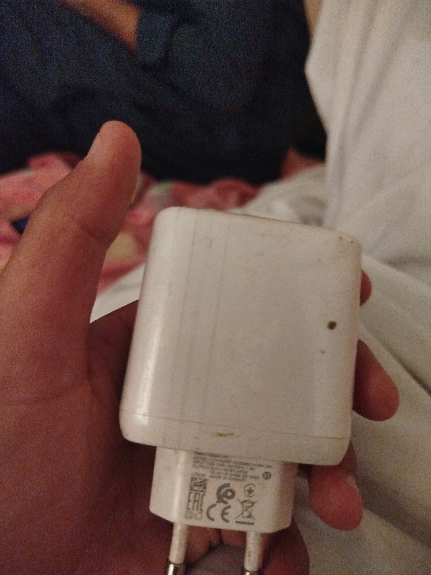 A close-up shot shows a person's hand holding a white USB power adapter. The adapter is rectangular with rounded edges and appears to be slightly dusty. Markings, including a logo with the letters "EAC" and a circular "CE" symbol, are visible on the adapter. The background consists of blurred dark fabric and white bedding. There is no discernible information to indicate the location within the city of Lassan, Pakistan.