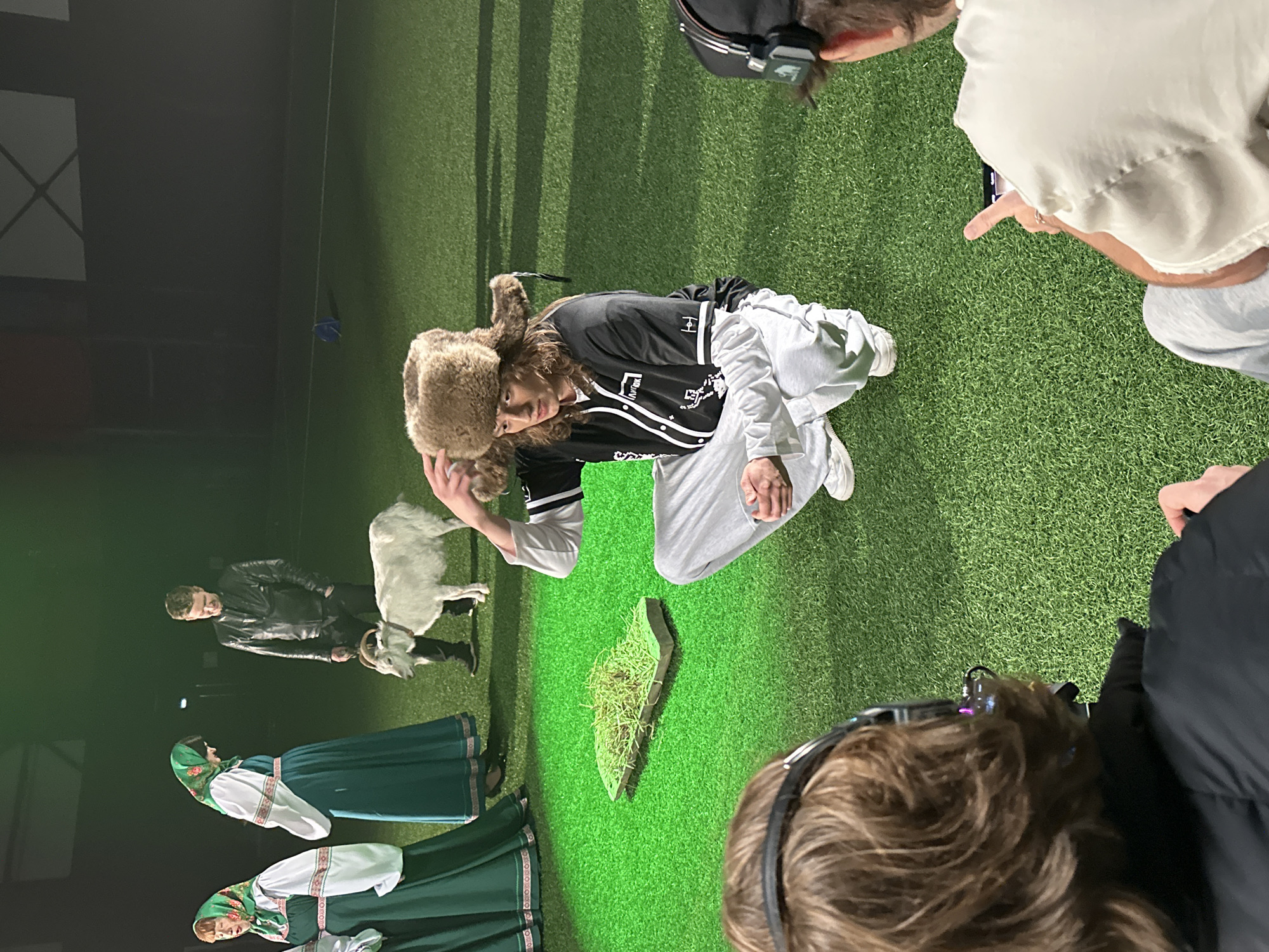 The image captures a scene in an indoor setting, possibly a theater or studio, in what appears to be a performance or staged event. A person wearing a furry hat and a black baseball jersey is kneeling on what looks like artificial grass. A white goat stands beside this person, and another person in a black leather jacket is holding the goat's leash.
Three people are dressed in traditional green and white folk costumes. An object that appears to be a small, rectangular platform covered in grass is present. Several people are observing and recording the scene with their mobile phones. The overall environment suggests a theatrical presentation or a performance piece. Based on the presence of traditional folk costumes, it is likely a Russian production, and the Balashikha location could be related to where the event took place.