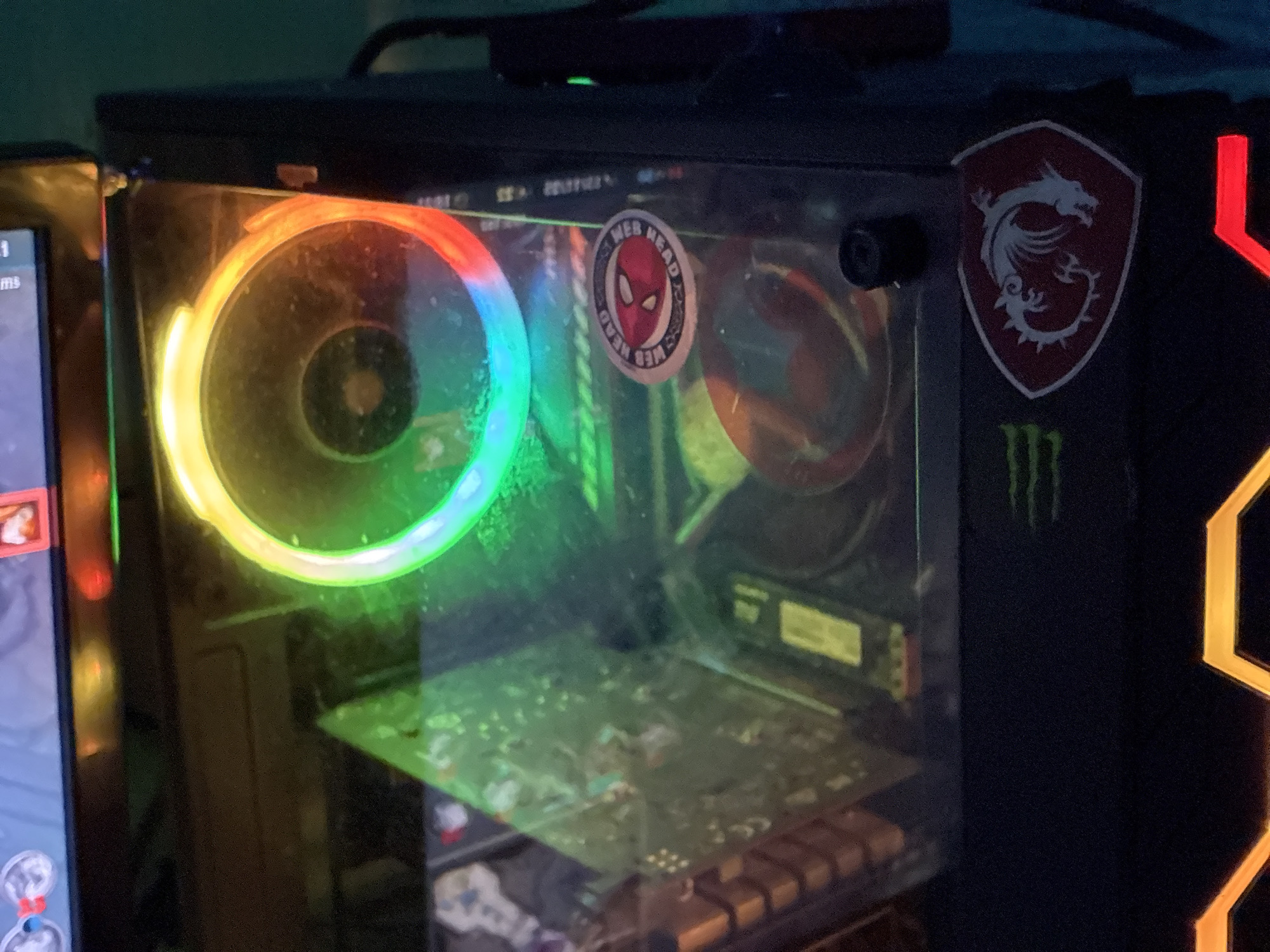 The image shows a partially visible computer tower with a clear side panel. Inside the tower, there's a circular fan with RGB lighting, a motherboard, and keyboard keys. A "Web Head" sticker is affixed to the side panel. On the outside of the case, a MSI logo sticker is visible, alongside some Monster Energy logo remnants and an orange geometric design. A monitor is visible to the left. The image does not contain sufficient information to identify the location as Elhovo, Bulgaria or any other location.