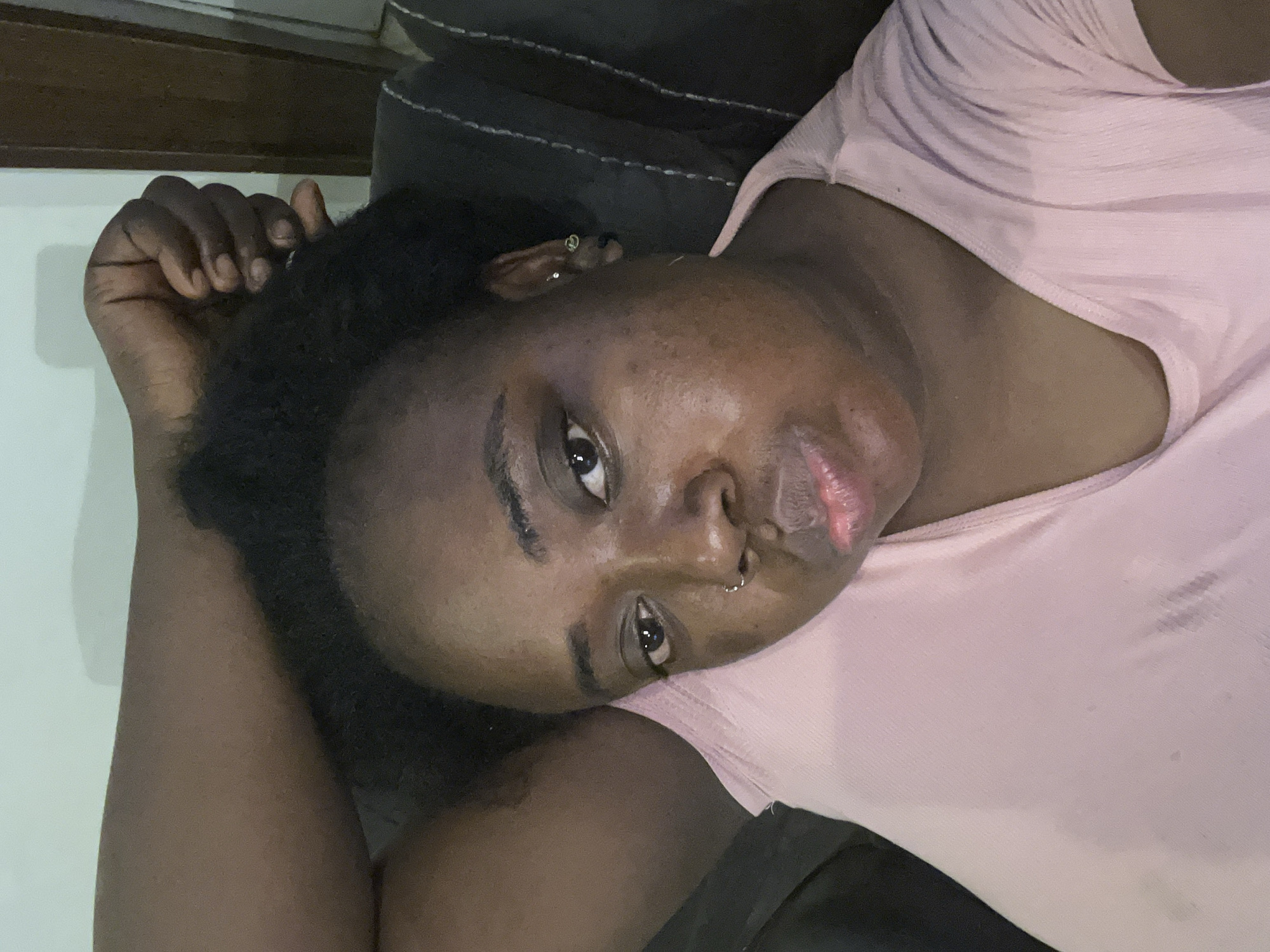 A dark-skinned woman with short, dark curly hair is lying down on what appears to be a dark-colored cushion or sofa. She is wearing a light pink t-shirt. She has a nose ring and earrings. Her right arm is raised near her head, with her hand near her hair. The location of the scene is unknown and cannot be confidently described as being in Jos, Nigeria from this photo. There are no definitive geographic or cultural markers visible in the image to confirm this location.