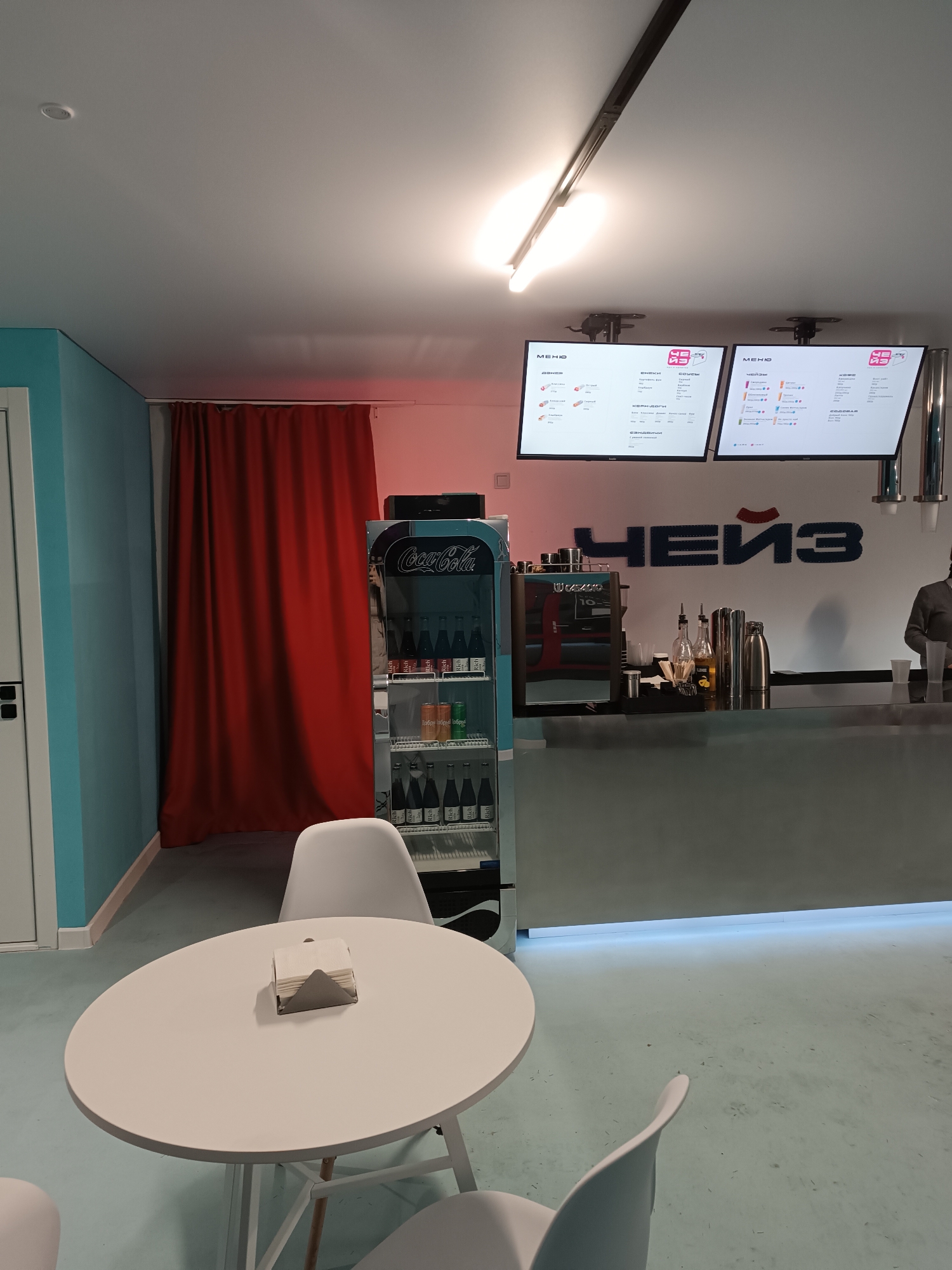 The scene is an interior shot of a modern cafe or counter-service establishment located in Unknown, Unknown. The room features a light blue wall on the left transitioning to a white wall on the right, with a large, floor-to-ceiling red curtain covering a section of the back wall. The floor is a light blue-grey, appearing to be a smooth, possibly polished, surface with minor scuffs and specks. The ceiling is white, equipped with a linear track light fixture and a single recessed light.
In the foreground, a white circular table stands centrally, topped with a silver-colored napkin holder containing white paper napkins. Partial views of two white chairs are visible around the table.
The midground reveals a silver, glass-fronted Coca-Cola branded refrigerator positioned to the right of the red curtain, stocked with multiple rows of various bottled beverages. To its right is a counter area. On the counter, from left to right, are a black and silver espresso machine, a tall silver container, two glass bottles with dispensers (one with a yellow label), a metal shaker, and a silver thermal carafe. Above the counter, a large blue logo or text reading "ЧЕЙЗ" (CHEIZ) is affixed to the white wall. Two illuminated digital menu screens displaying text and colorful graphics in Cyrillic are mounted above the counter.
A single individual, visible from the torso upwards, stands behind the right side of the counter. The person wears a dark grey long-sleeved top, with a clear plastic cup visible near their hand, suggesting active work or preparation. No specific interactions are depicted. The ambient lighting is bright, supplemented by a subtle red hue emanating from behind the red curtain.