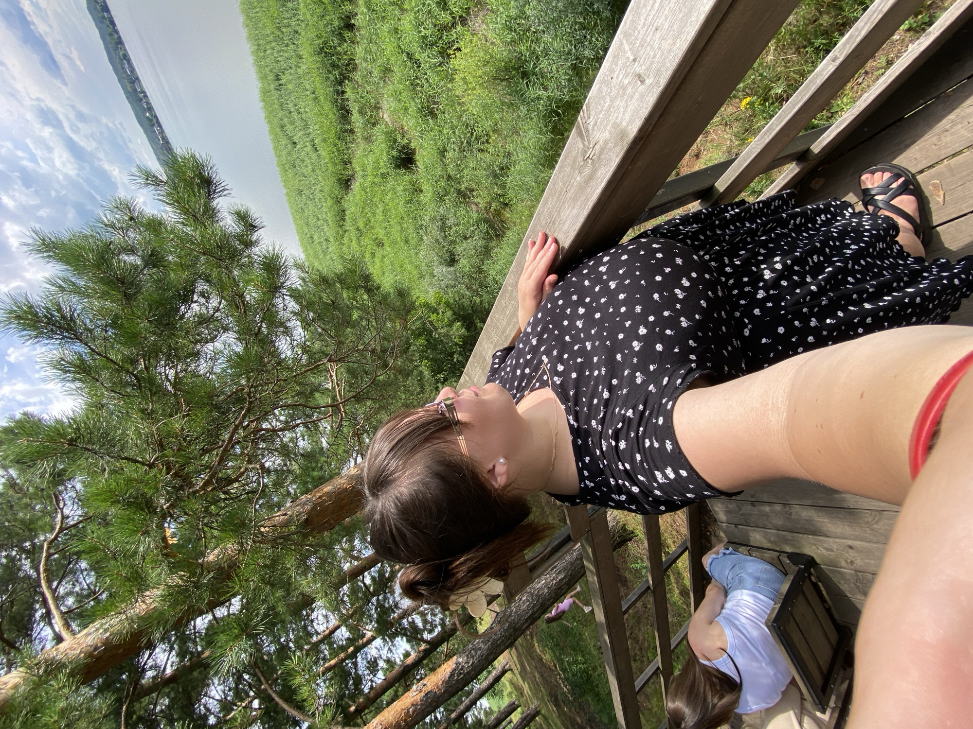 This media file features two individuals on a wooden deck overlooking a lush green landscape and a body of water. The primary subject, a woman wearing a black and white floral dress and sandals, is leaning on a wooden railing. She is wearing glasses and her hair is pulled back. Her hand rests on the railing, and her gaze is directed towards the scenery.
In the background, another person, possibly a child, is sitting on a low wooden bench or stool. They are wearing a white top and blue shorts or jeans and their long brown hair is visible.
The scene appears to be set in a natural environment, possibly a park or a lakeside area. The greenery suggests it is daytime, and the lighting indicates it's likely sunny. The presence of a pine tree in the foreground adds to the natural setting. The overall mood seems relaxed and peaceful, with the subjects enjoying the view. The location is identified as Votkinsk, Russia. There is no visible text in the image.