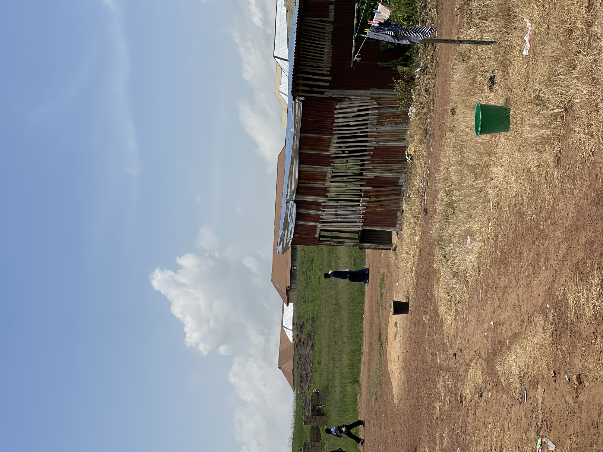 The image depicts a scene in Durumi, Nigeria, under a bright, partly cloudy sky, suggesting it is daytime. The setting appears to be a residential area characterized by informal housing with corrugated metal roofs and walls. Several people are visible. In the foreground, a person in dark clothing stands near a patch of dry grass and dirt. Further back, another person in dark blue clothing is seen near a grassy area adjacent to the buildings. There are two more figures in the distance, one seemingly walking.
The buildings themselves show signs of wear and tear, with sections of corrugated metal exposed and wooden slats used as makeshift supports or coverings. Laundry is hung on a line outside one of the structures, with several items of clothing visible. A bright green bucket sits in the dry grass, and a dark rectangular object, possibly a bin or a small structure, is also present in the scene. The ground is a mixture of dry, yellowish grass and reddish-brown dirt, with some scattered debris. The overall impression is of a lived-in, possibly developing, community. No visible text is present in the image.