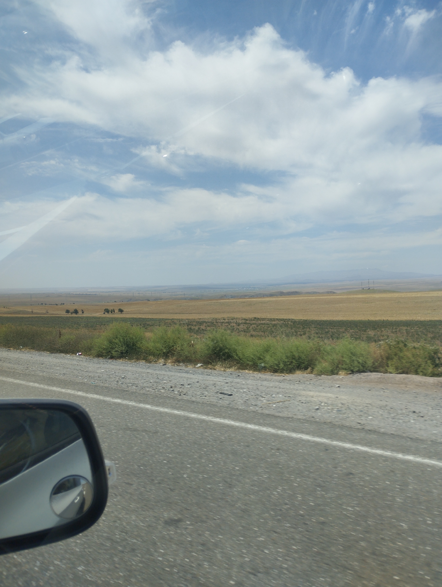The image captures a roadside view from a vehicle, likely in a rural or semi-rural area. The foreground shows a section of asphalt road with a white line marking the lane. To the left, a side-view mirror of the vehicle is visible, reflecting a distorted view of the road and sky.
The middle ground features a dry, grassy landscape with scattered patches of green shrubs and vegetation along the edge of the road. In the distance, a vast, flat expanse of dry fields stretches towards the horizon. A few clusters of trees are sparsely dotted across this landscape, and in the far distance, faint outlines of hills and mountains can be seen. The sky is predominantly blue with scattered white clouds, some of which appear wispy and elongated.
The scene appears to be a snapshot of a journey through a landscape characterized by open fields and a dry climate, typical of many regions in Uzbekistan, including areas around Sangzar. There are no people or specific identifiable structures that would pinpoint the exact location within Sangzar without further context. The overall impression is one of a tranquil, expansive rural environment under a partly cloudy sky.