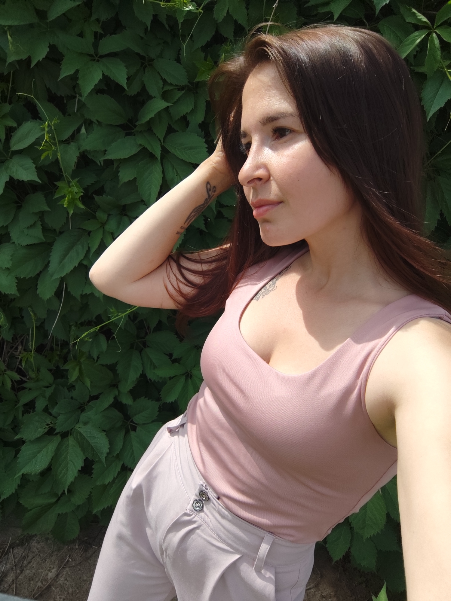 A woman stands against a dense wall of green foliage, likely climbing vines, in Фоки, Russia. She is fair-skinned with long brown hair. She wears a mauve tank top and matching slacks. Her right arm is raised behind her head, and a tattoo is visible on the inside of her left forearm. Another tattoo is visible at the upper part of her chest. The photo appears to be a selfie, with the camera angle from slightly below and to the side. Sunlight highlights her face and upper body, while casting shadows within the leaves.